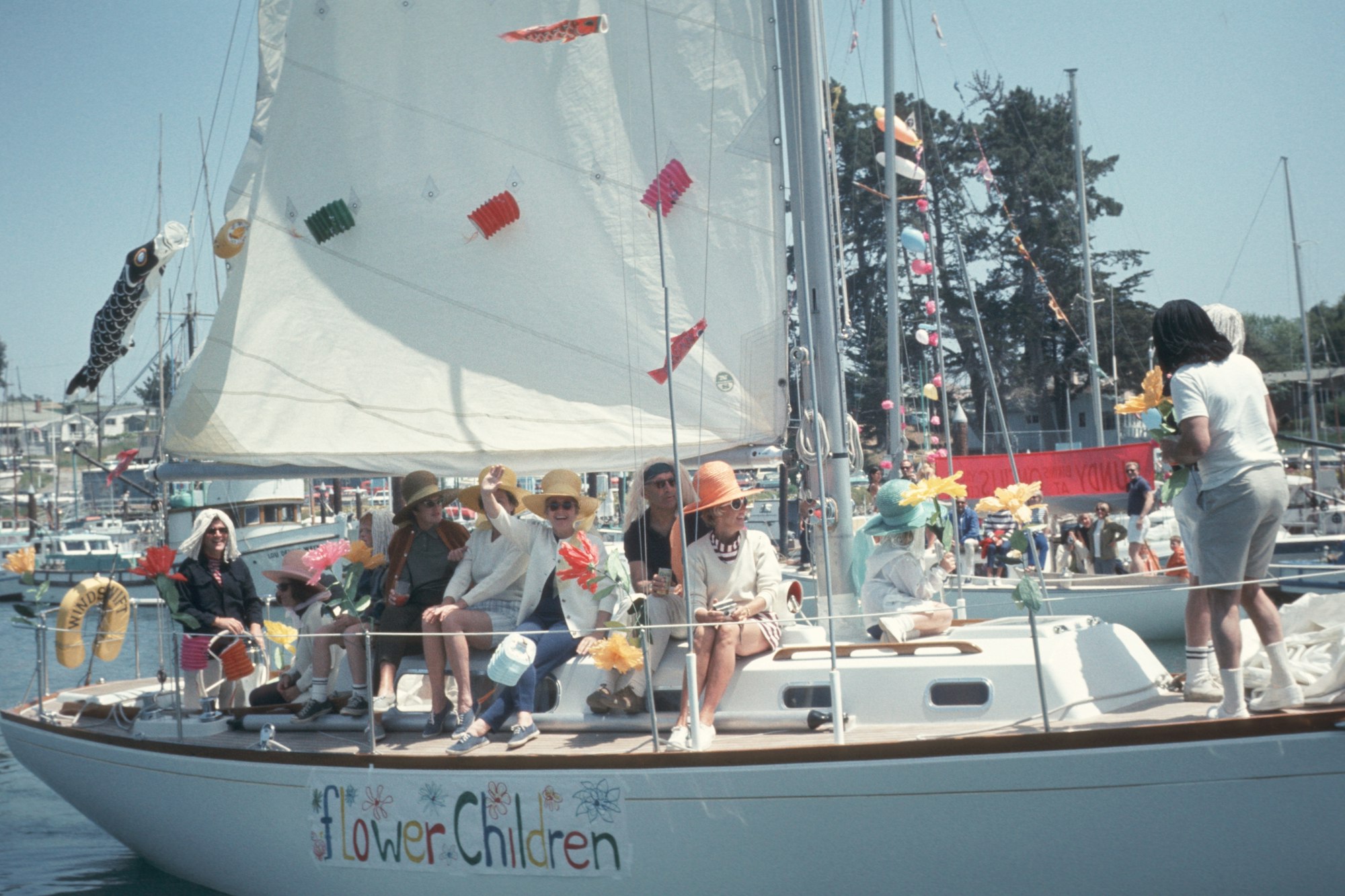 A group of people on a decorated sailboat named "Flower Children," waving and enjoying the day.