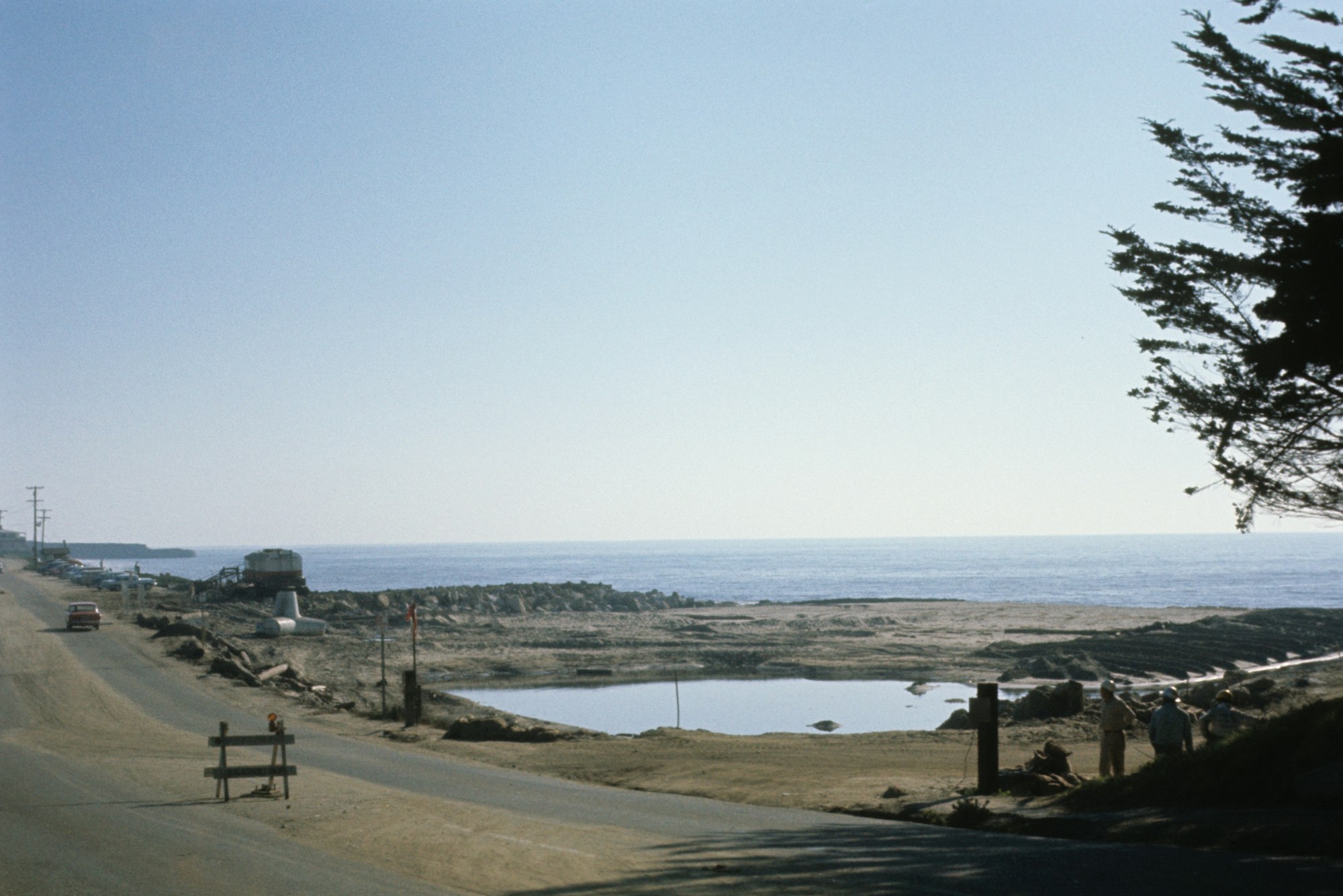 Coastal road with vehicles, ocean view, people, trees, and a clear sky.