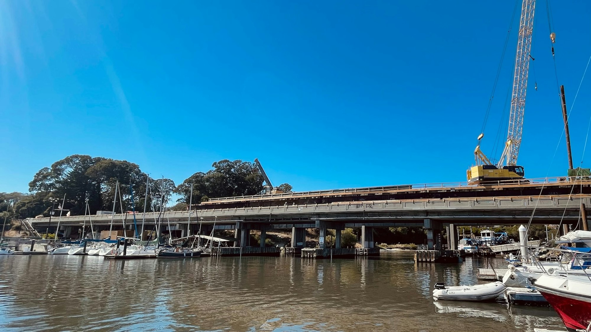Boats docked near a bridge under construction, with cranes and clear blue skies.