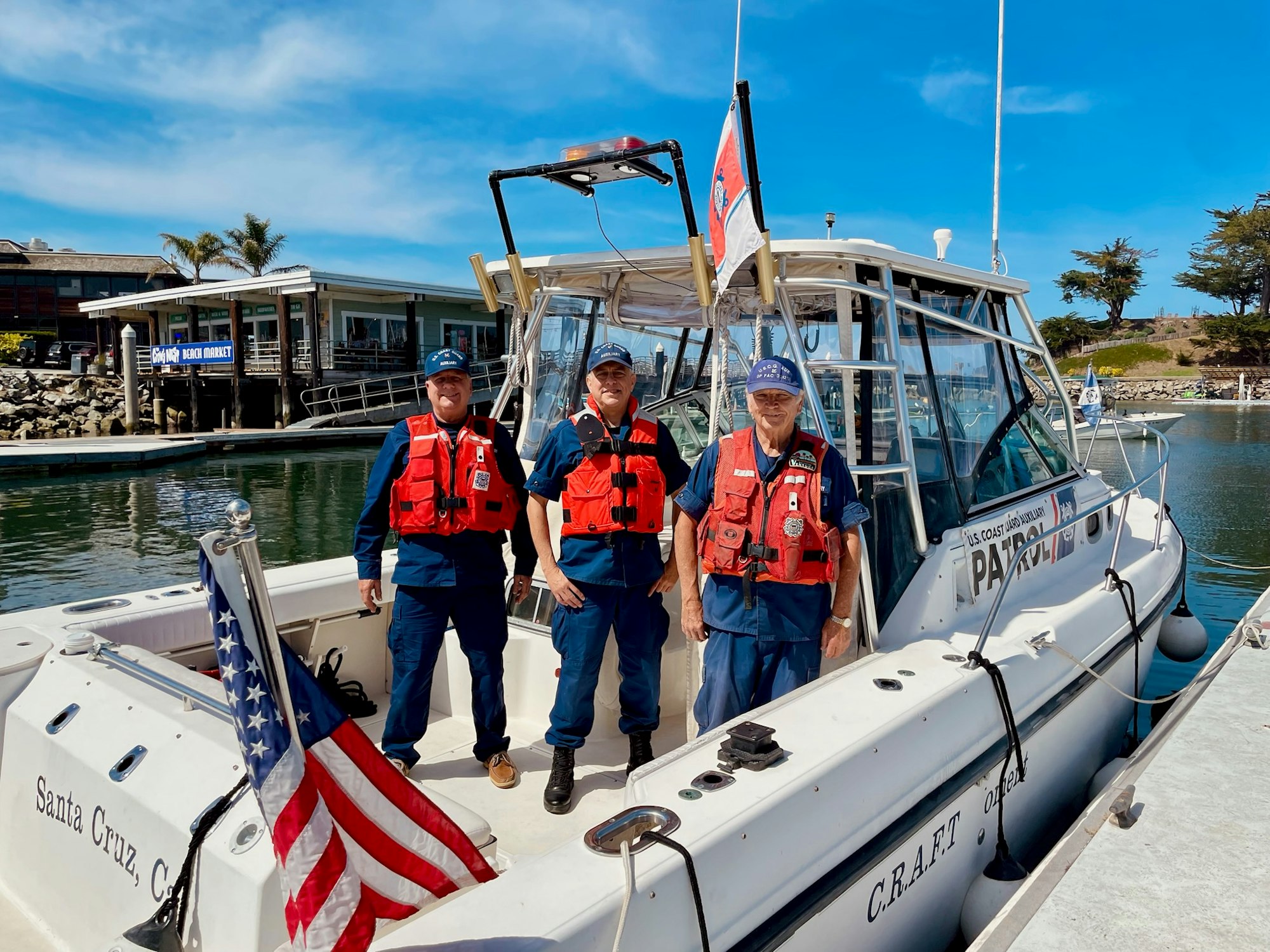 Three members of the U.S. Coast Guard stand on a boat at a marina, with an American flag and a clear blue sky in the background.