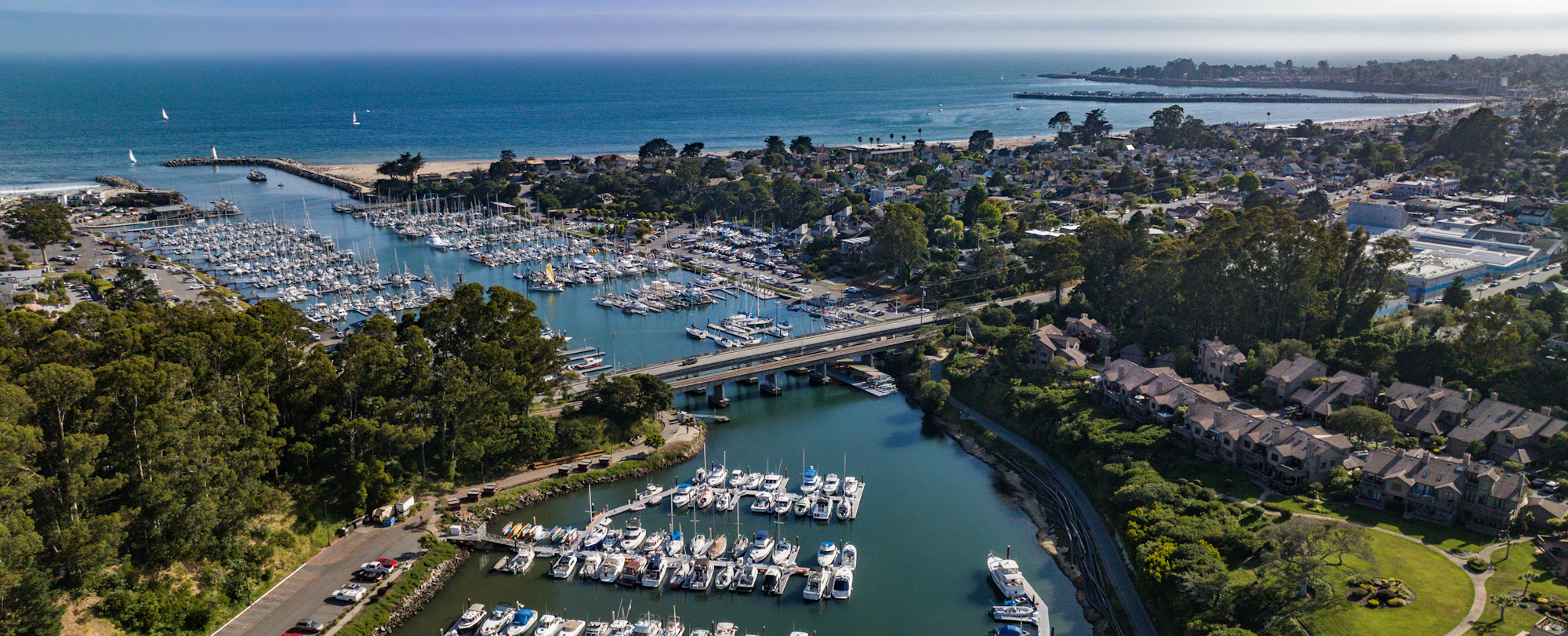 Aerial view of a marina with boats, nearby greenery, residential area, and ocean in the background.