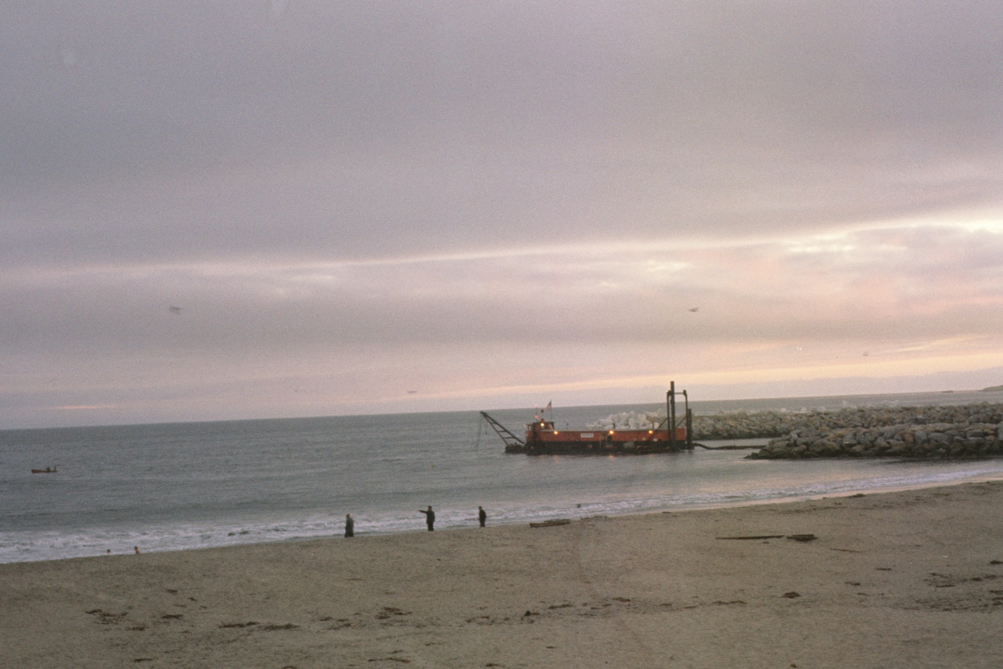Beach at dusk with people, a ship near rocks, calm sea, cloudy sky.