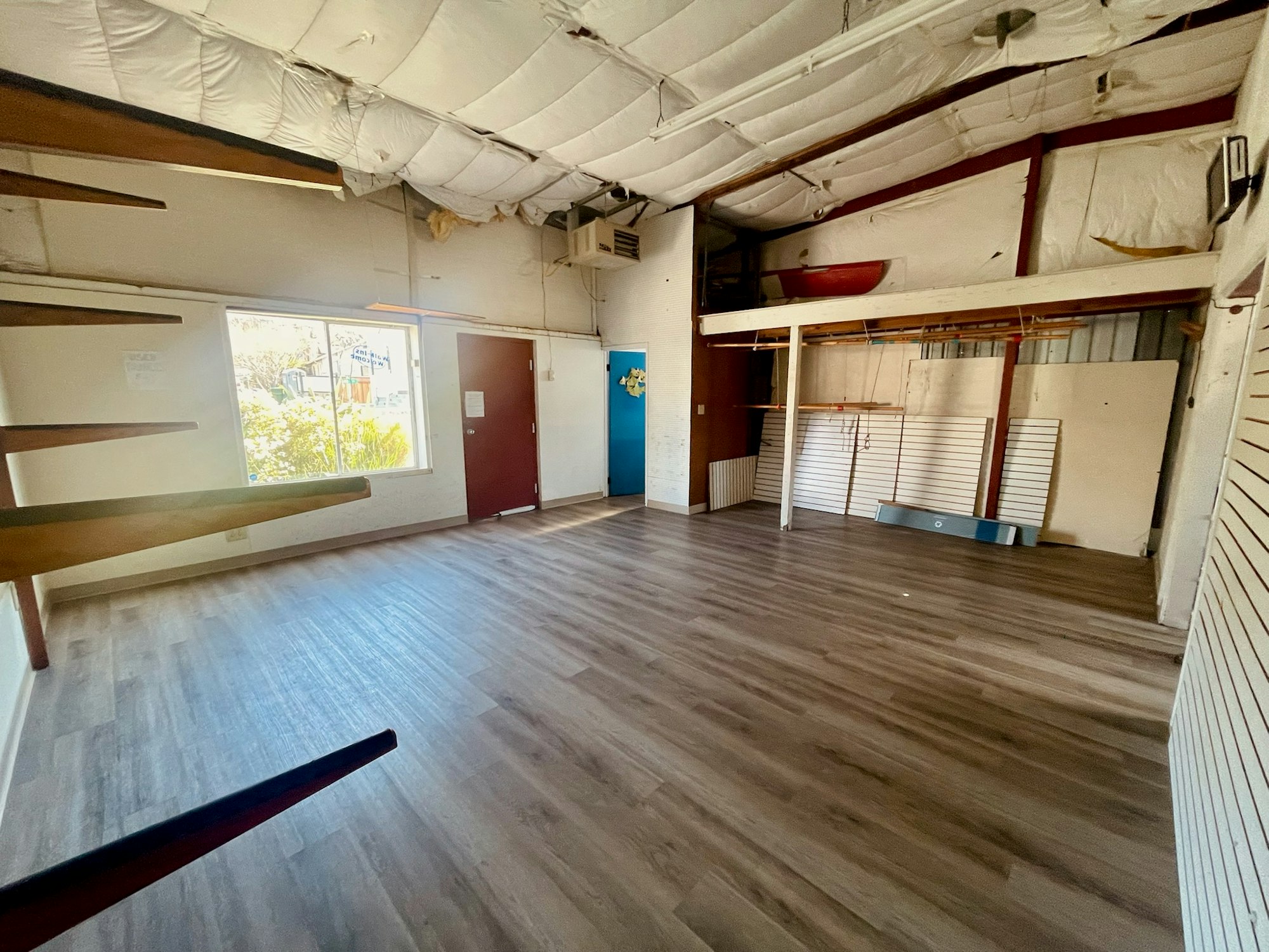 An empty interior space with wooden flooring, shelves, a large window, and minimal furniture or decorations.