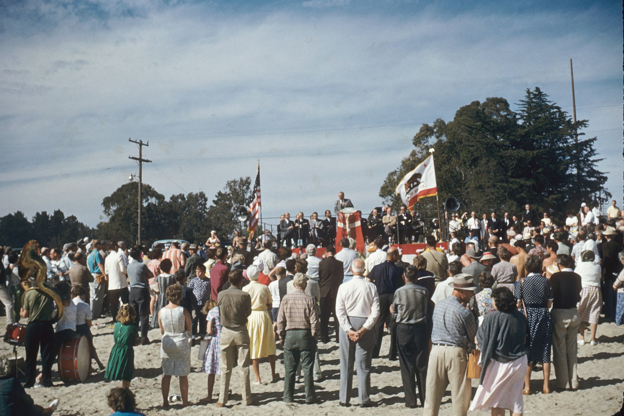 A crowd gathered for an outdoor event, with a stage and flags, possibly a political rally or community meeting.