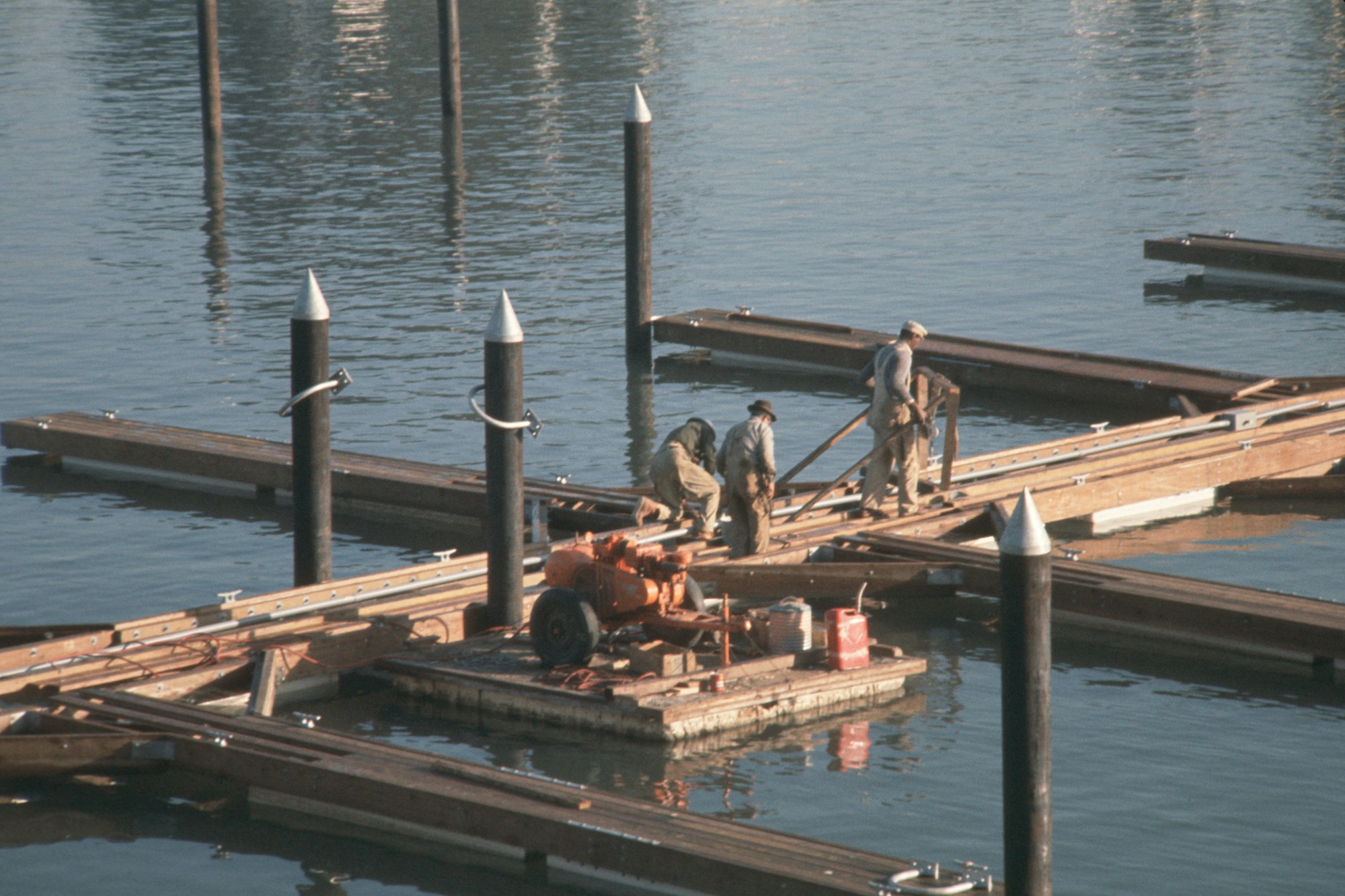 Construction workers repairing or building a wooden dock on calm water, with tools and materials around.