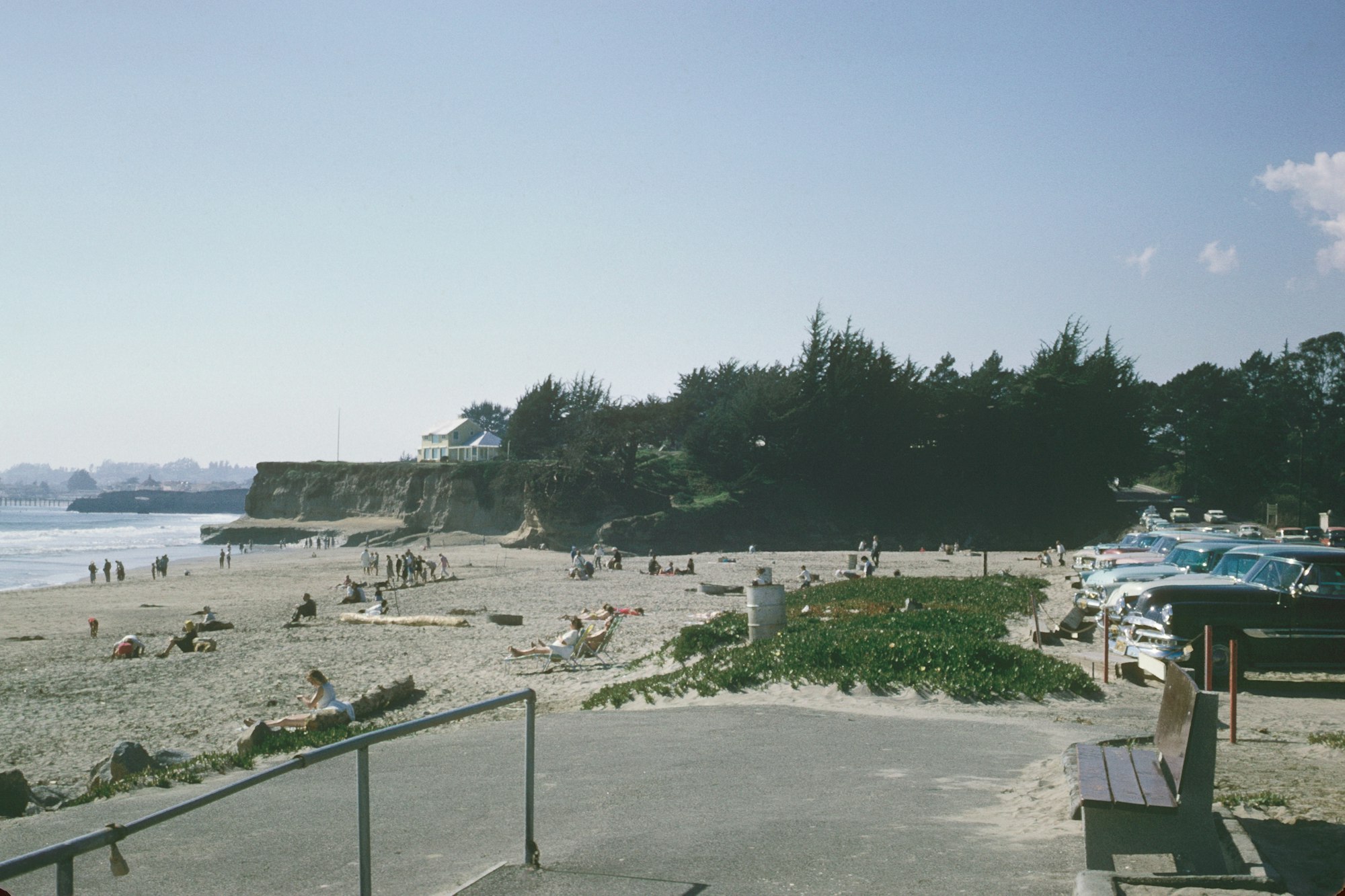 A sunny beach with people, cliffs, trees, vintage cars, and a blue house. Vintage vibe.