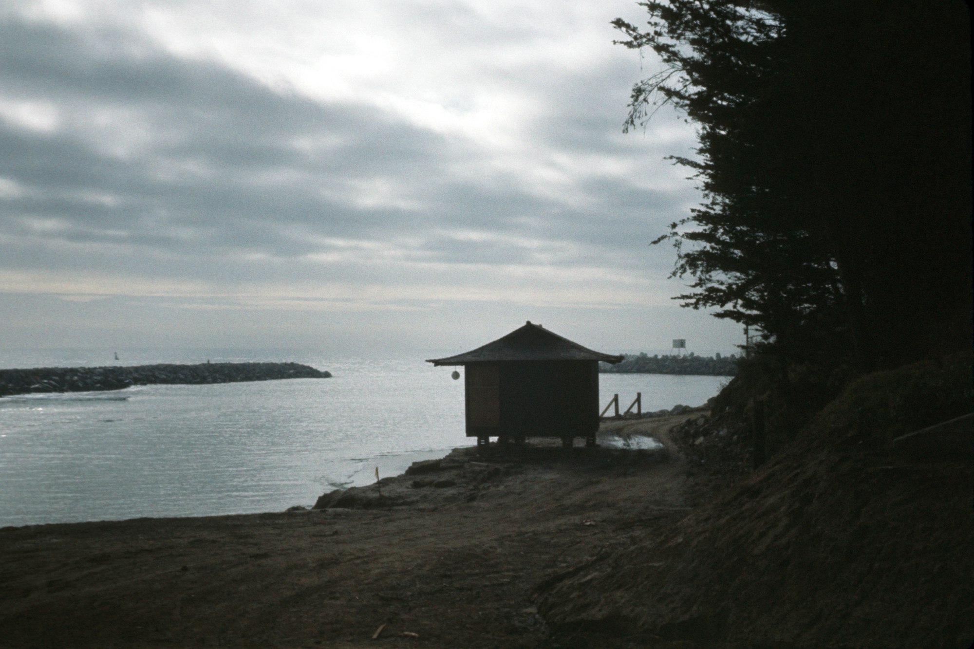 A small hut by the sea with cloudy skies and distant city skyline.
