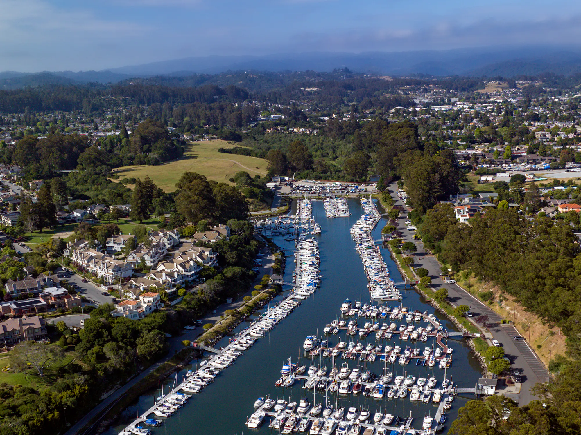 Aerial view of a marina filled with yachts, surrounded by green hills and residential areas, under a clear blue sky.