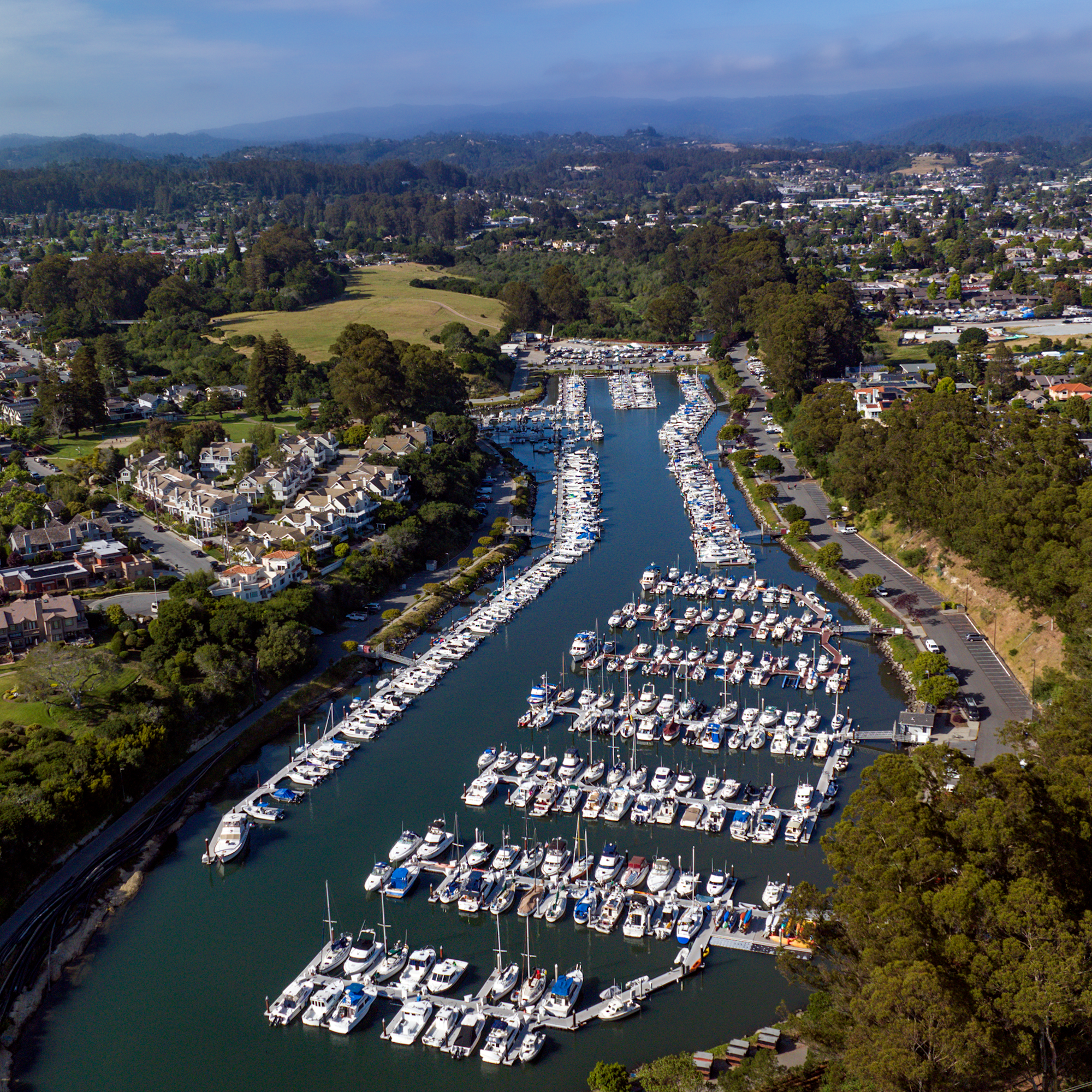 Aerial view of a marina filled with yachts, surrounded by green hills and residential areas, under a clear blue sky.
