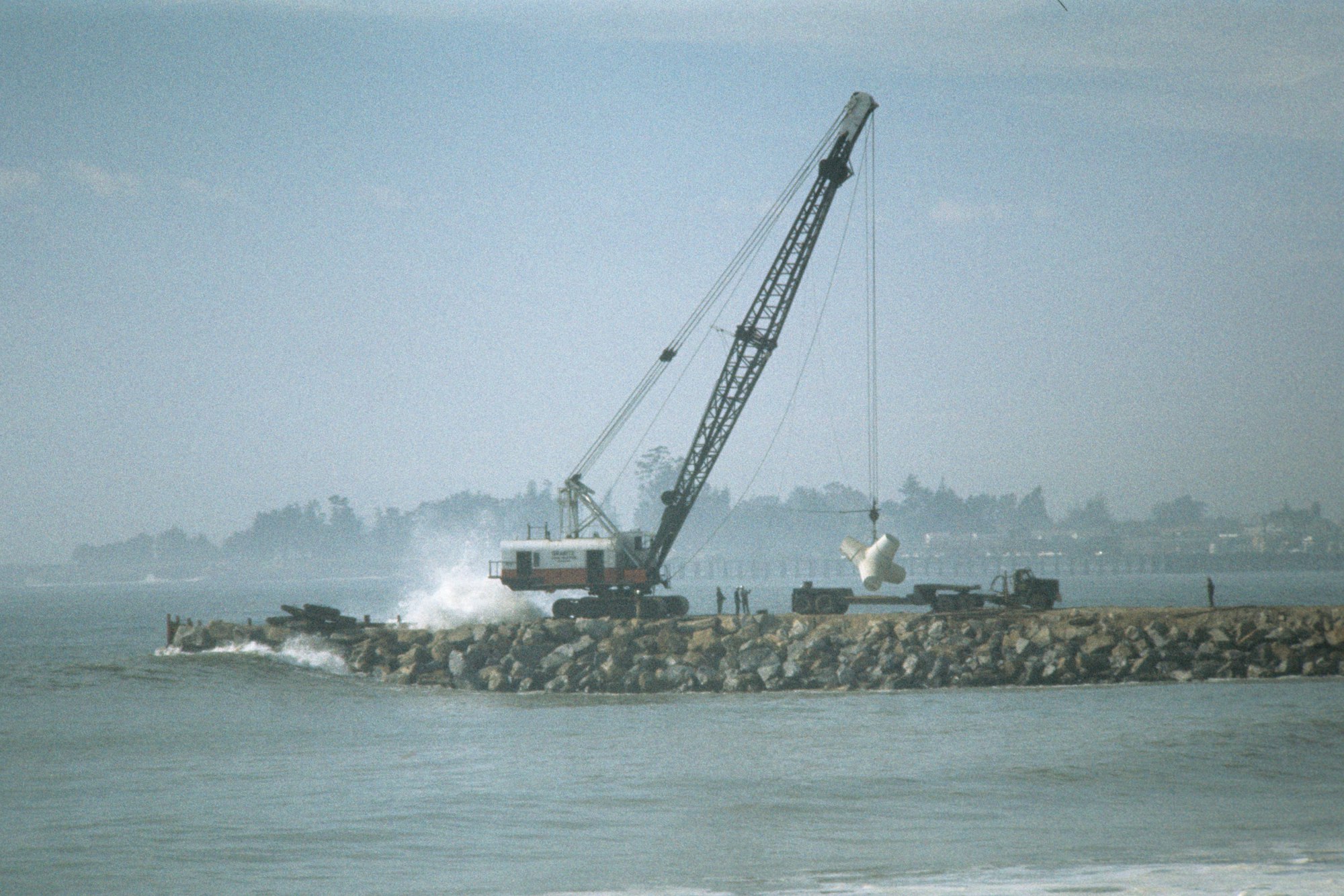 A crane on a breakwater lifting material, with waves crashing and trees in the background.