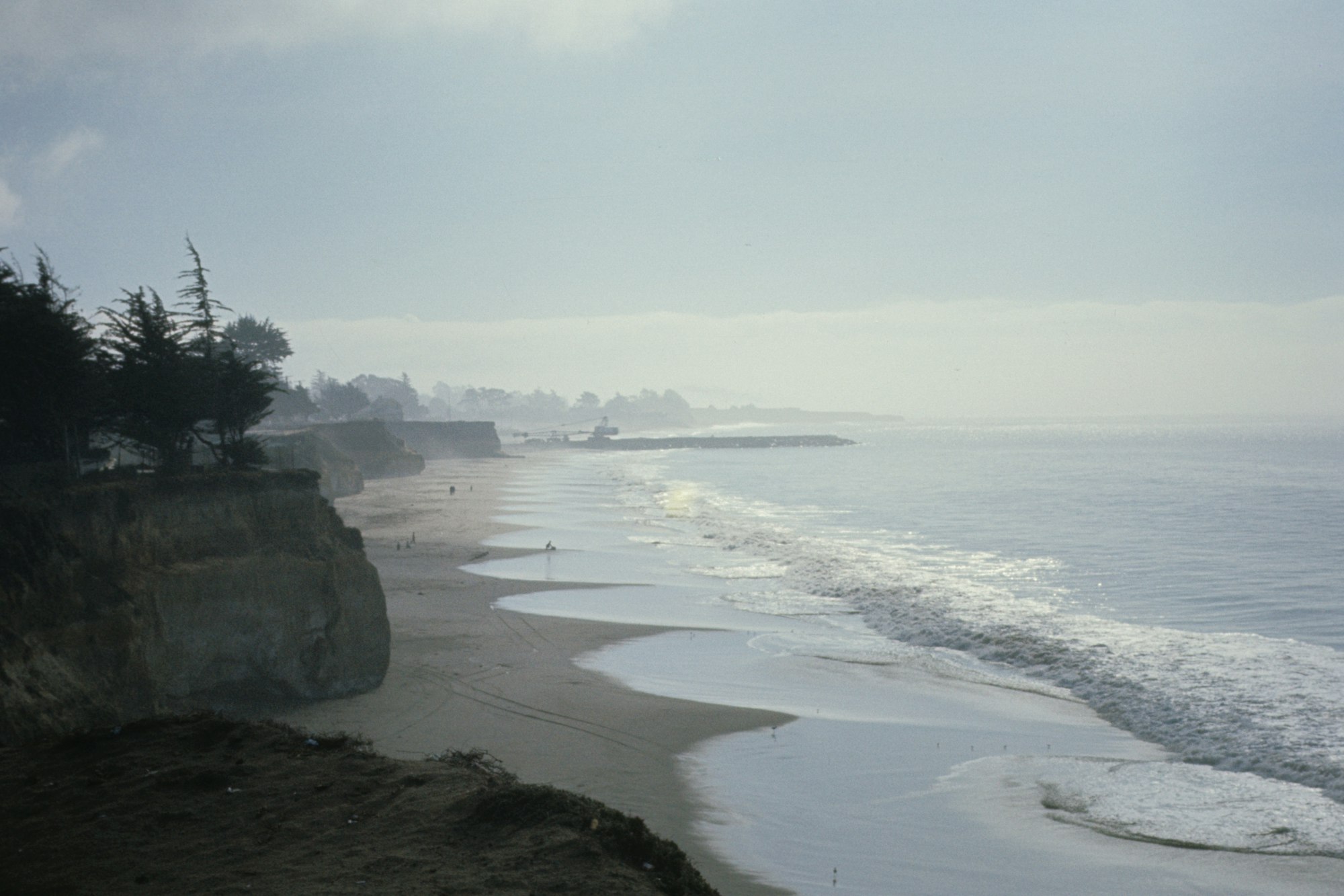 A coastline with cliffs, a sandy beach, waves, people at a distance, and overcast skies.