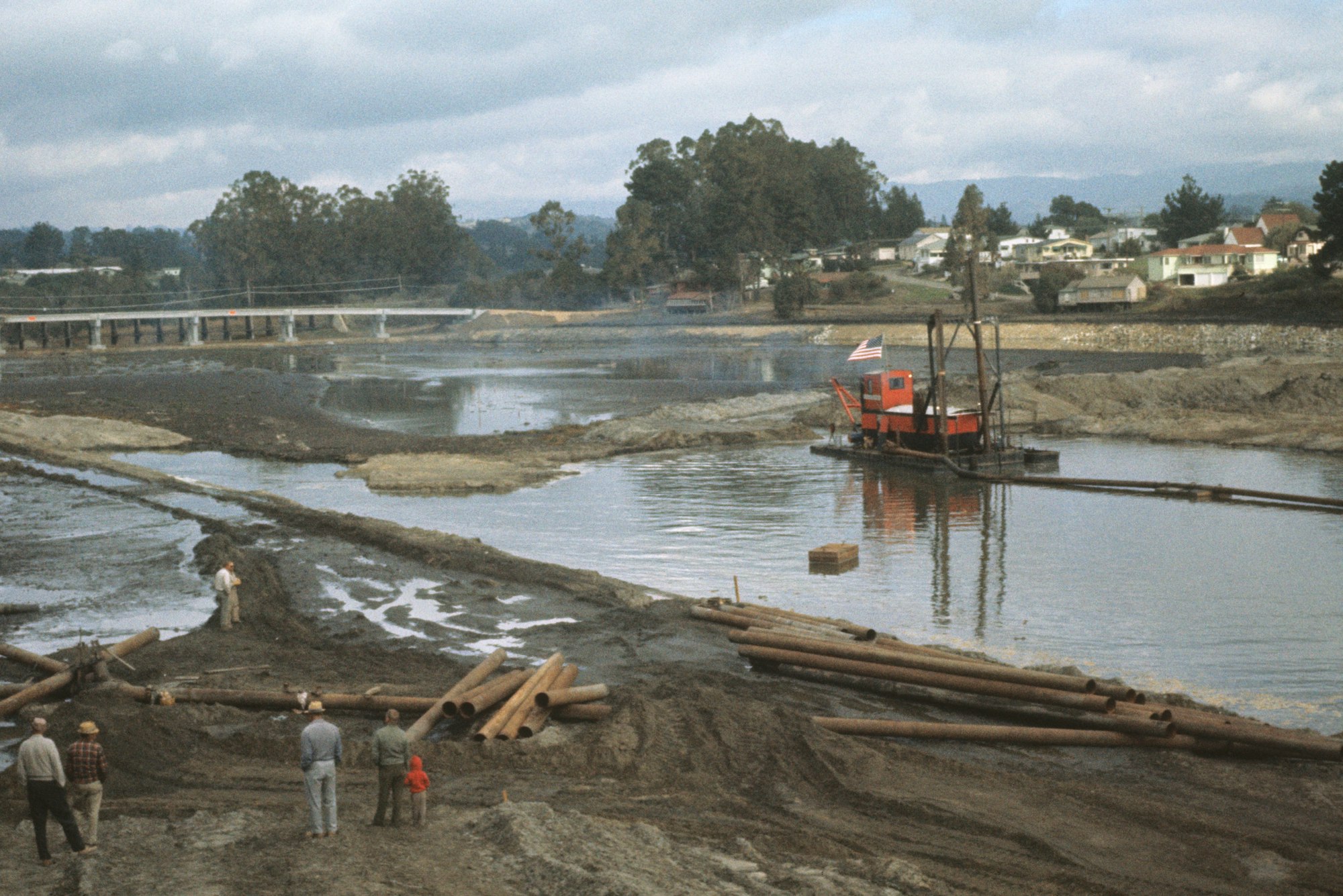 Dredging operations with a crane on a barge, people observing, pipes on shore, bridge and houses in the background.