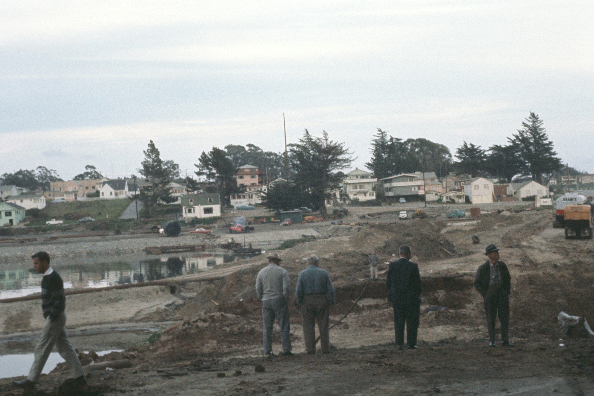 Men observing a construction site near a residential area with vehicles and equipment, and a dog in the foreground.