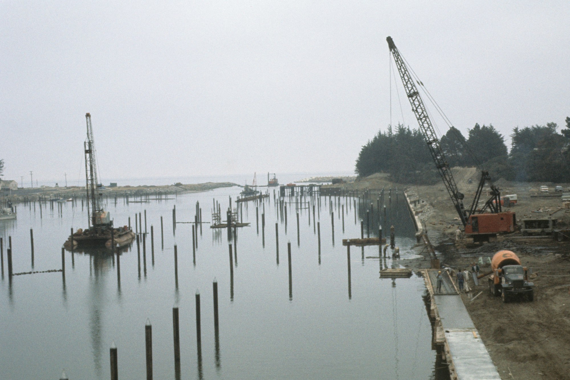 Construction site at a waterfront with cranes, piles, and machinery.