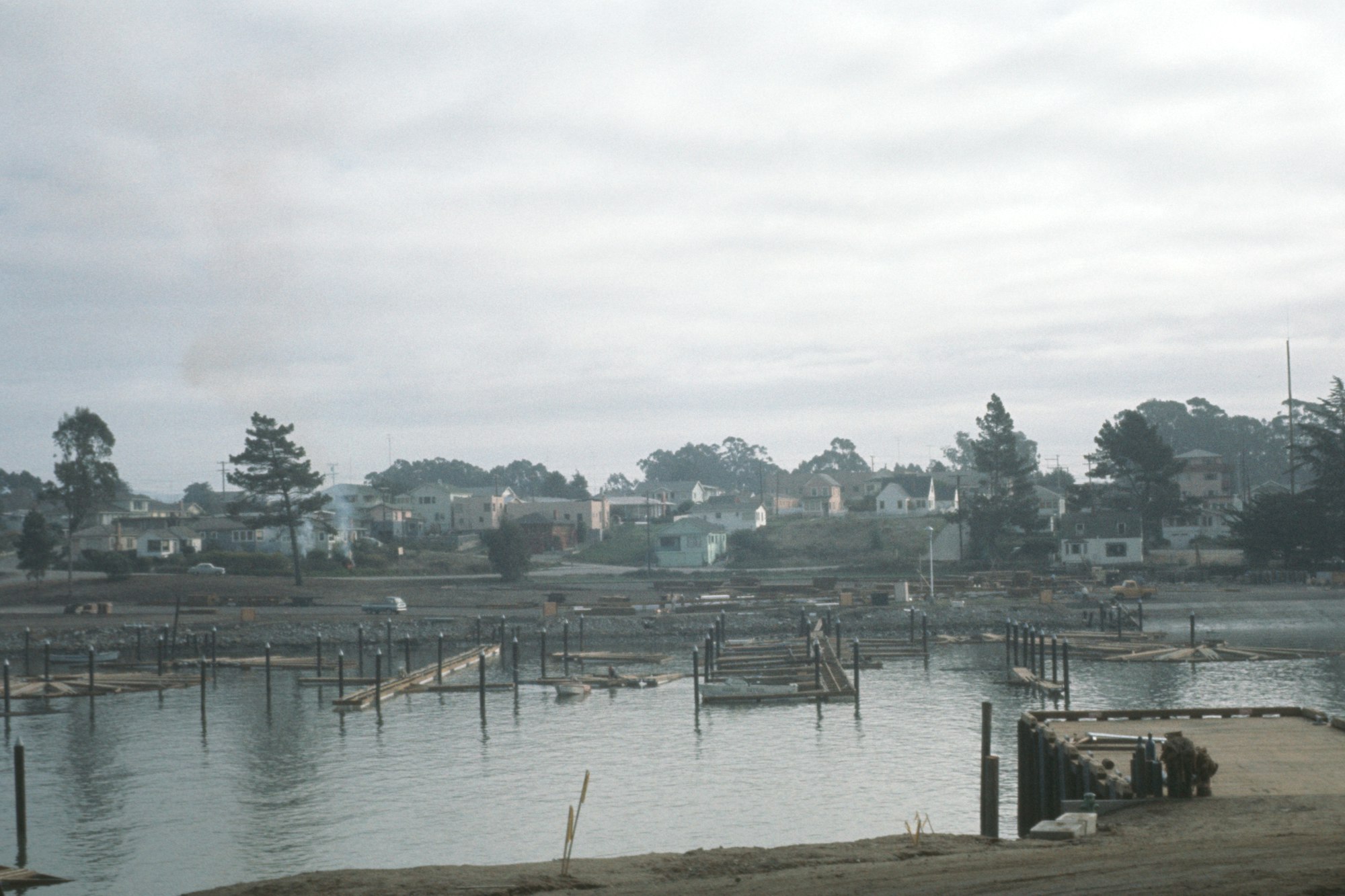 A waterfront construction area with wooden docks and residential buildings in the background under an overcast sky.