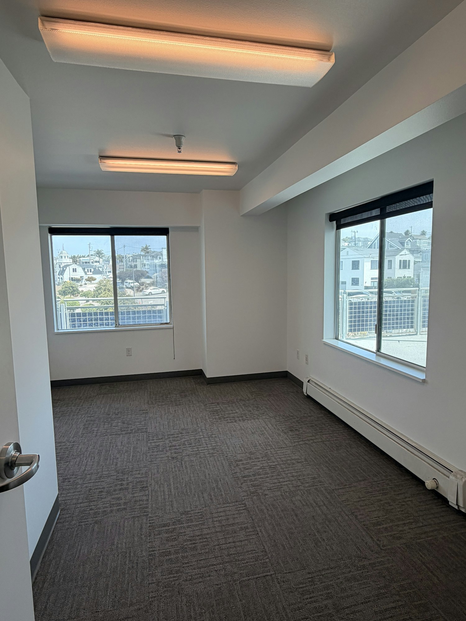 The image shows a bright, empty room with large windows and modern lighting fixtures, carpeted flooring, and an urban view.