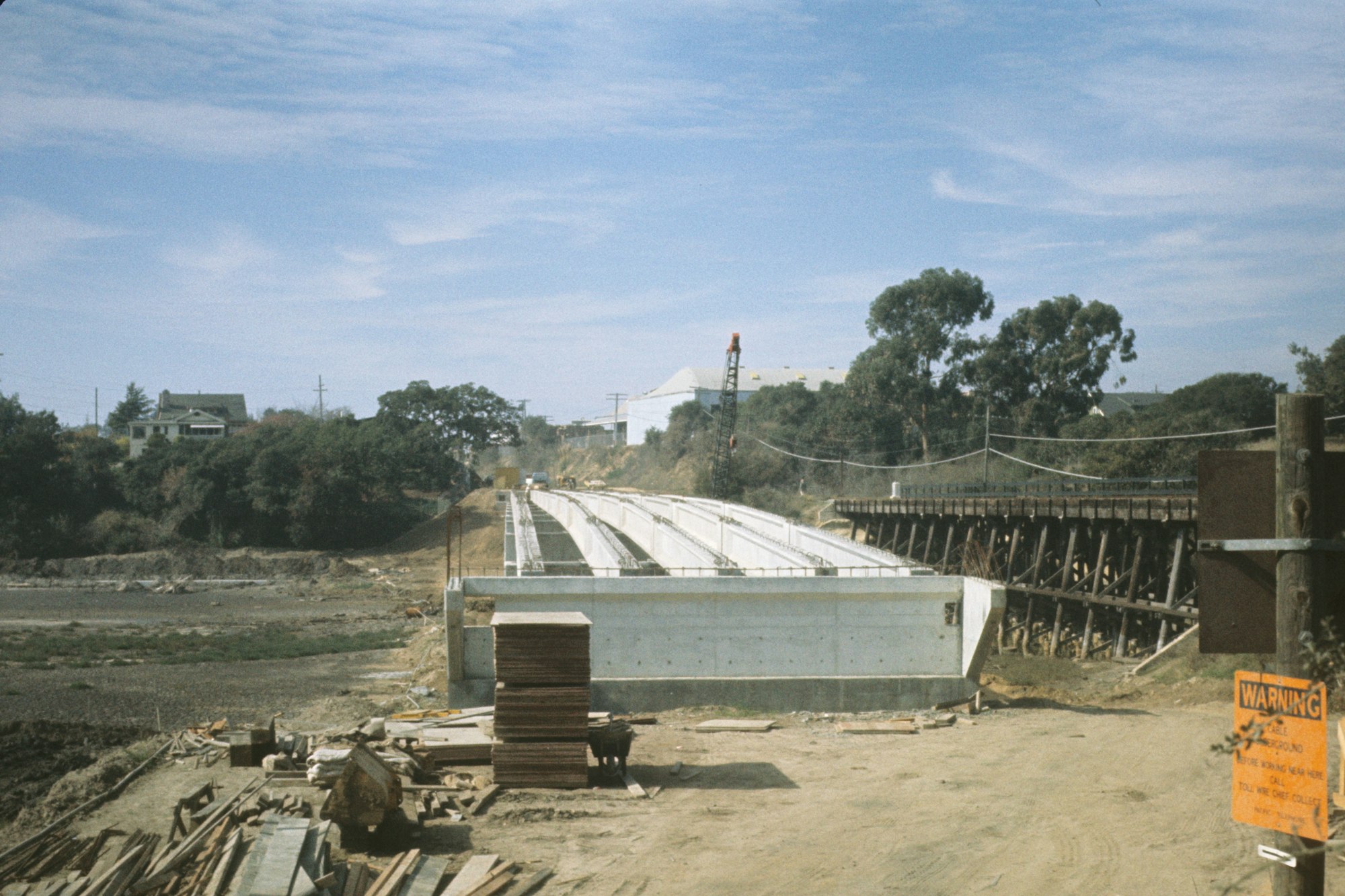 A construction site showing new concrete infrastructure next to an older wooden structure, equipment scattered around, and warning signs.