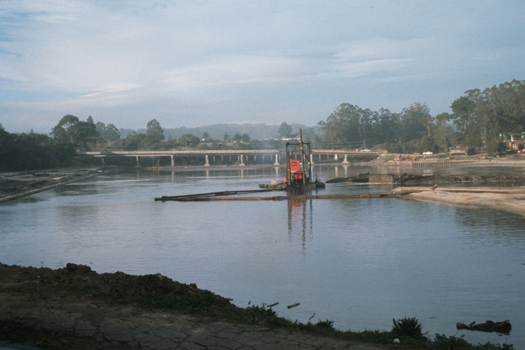 A dredger in a river near a bridge with trees in the background and a clear blue sky.