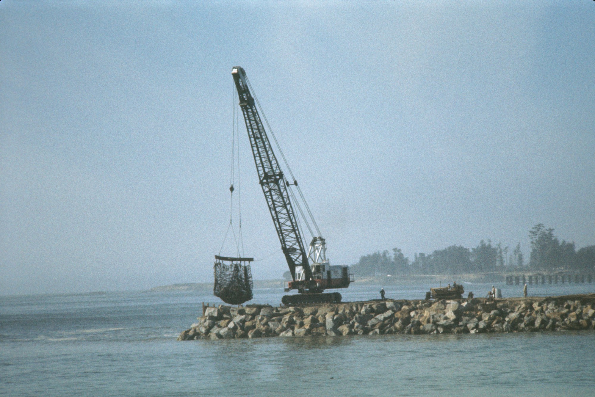Crane lifting materials at a coastal construction site with rock barrier.