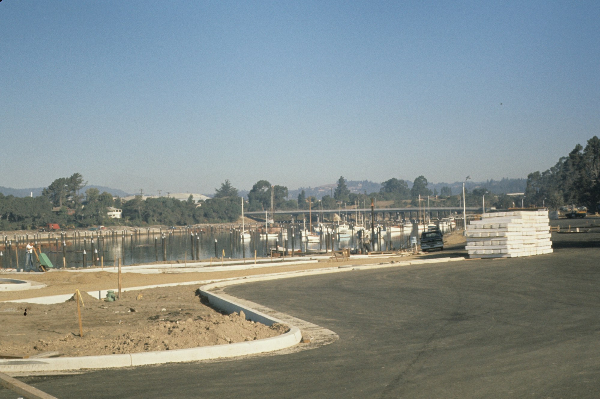 New road by a waterside construction area with concrete piles, clear sky, and trees in the background.