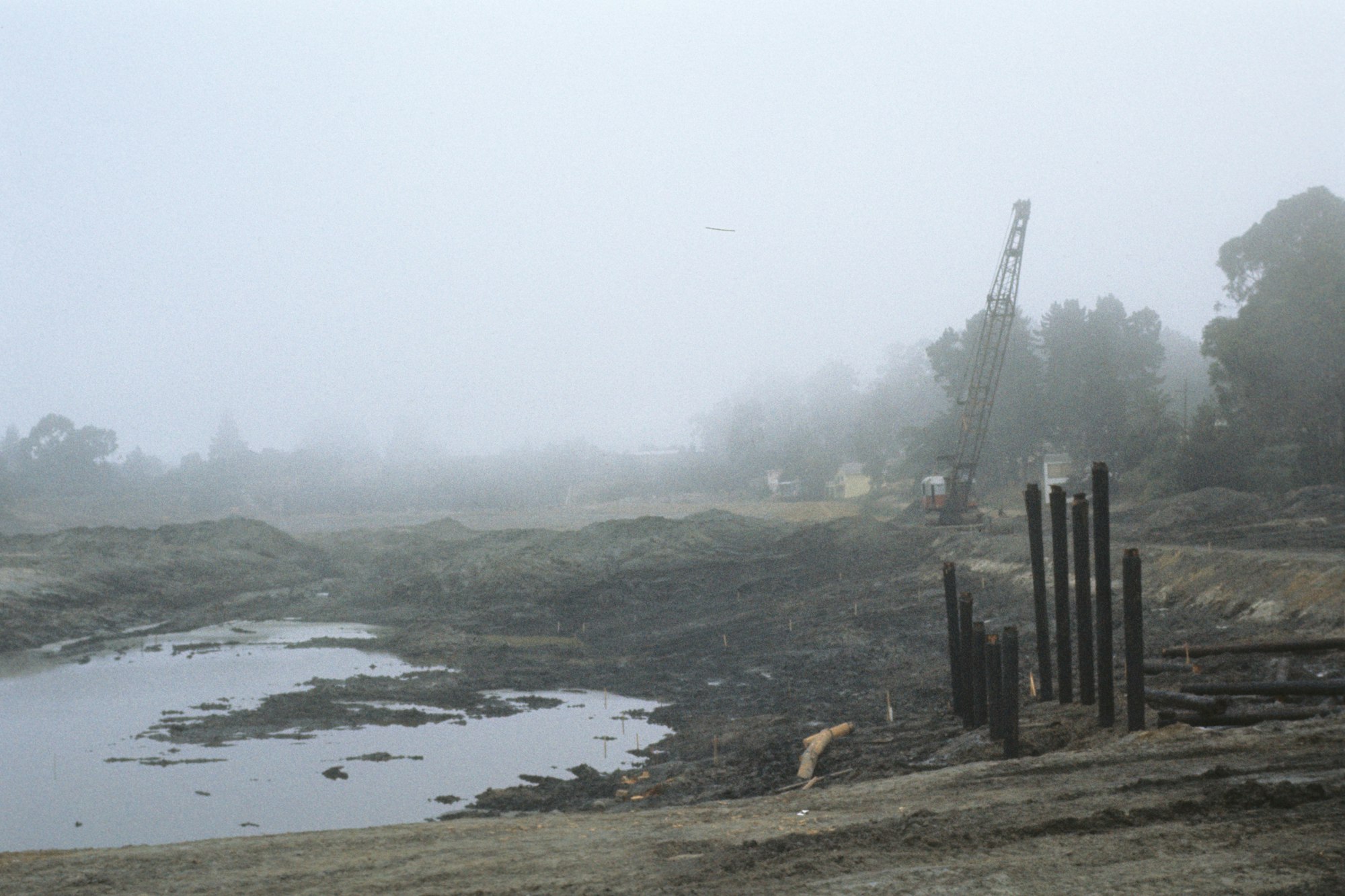 Foggy construction site with standing water, excavated earth, crane, and piles.