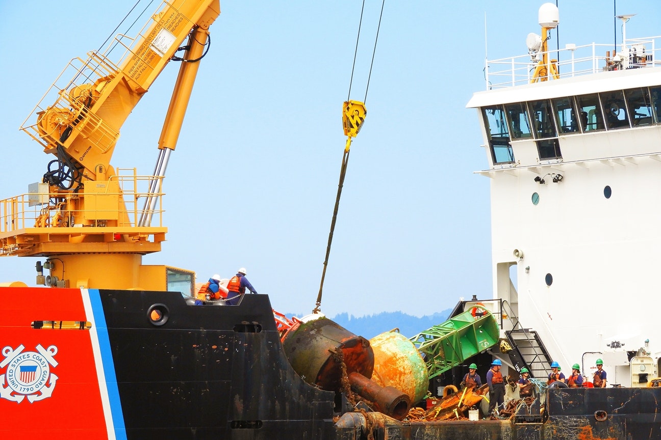 A crane lifting rusty equipment onto a ship with the US Coast Guard emblem, workers in safety gear are present.