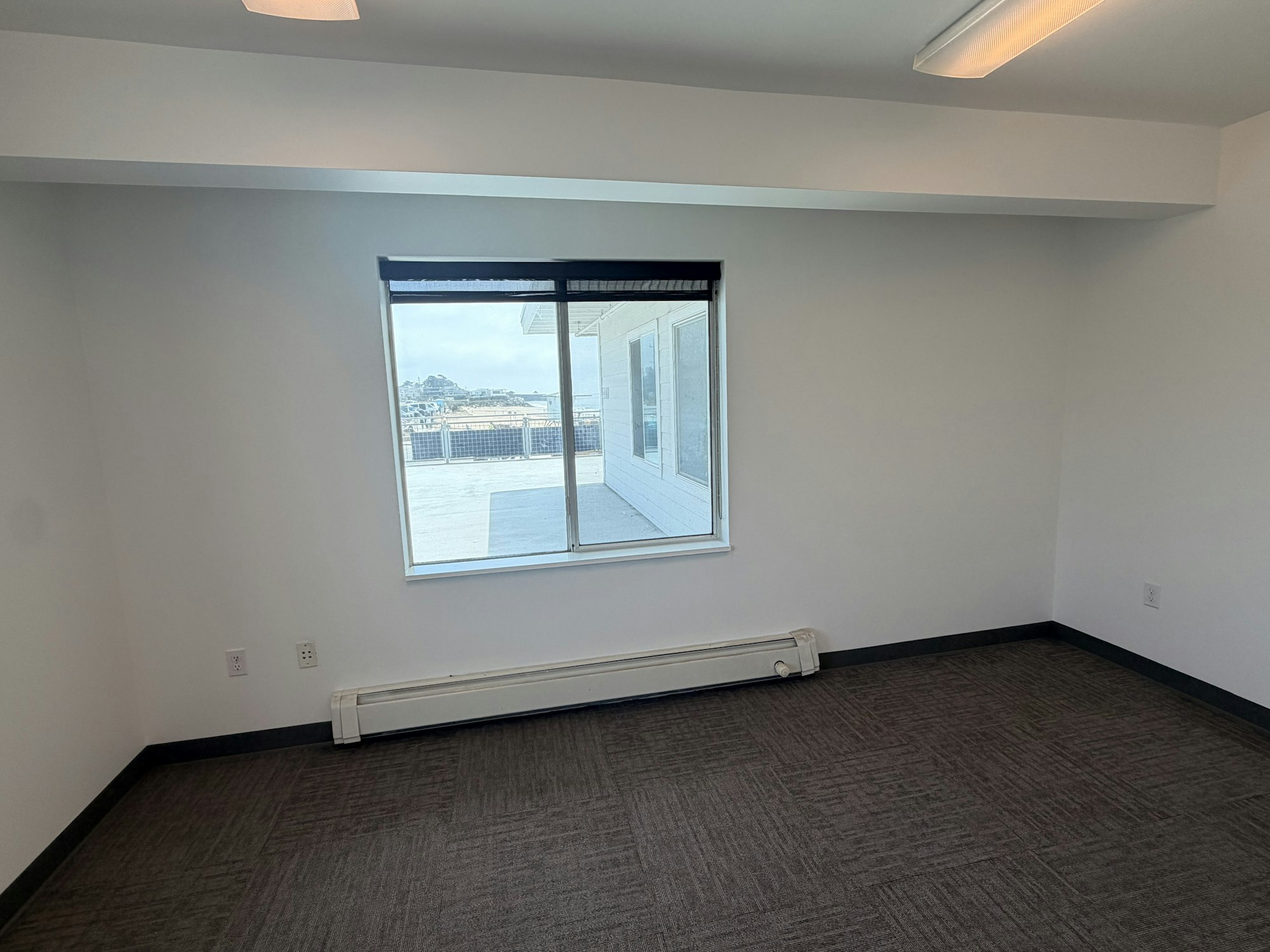 The image depicts a simple, empty room with a large window, light walls, and carpeting.