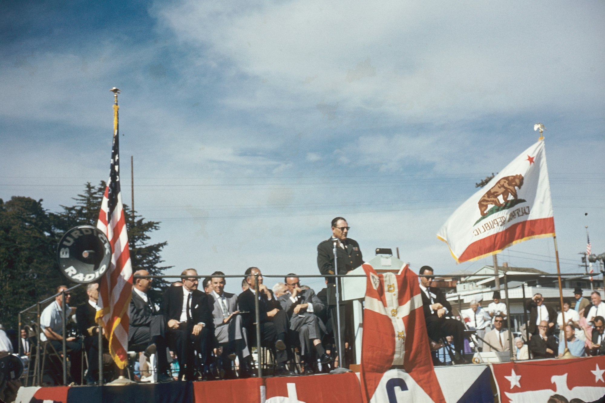 A man speaks at a podium with flags, people seated behind him, outdoor setting, vintage appearance.