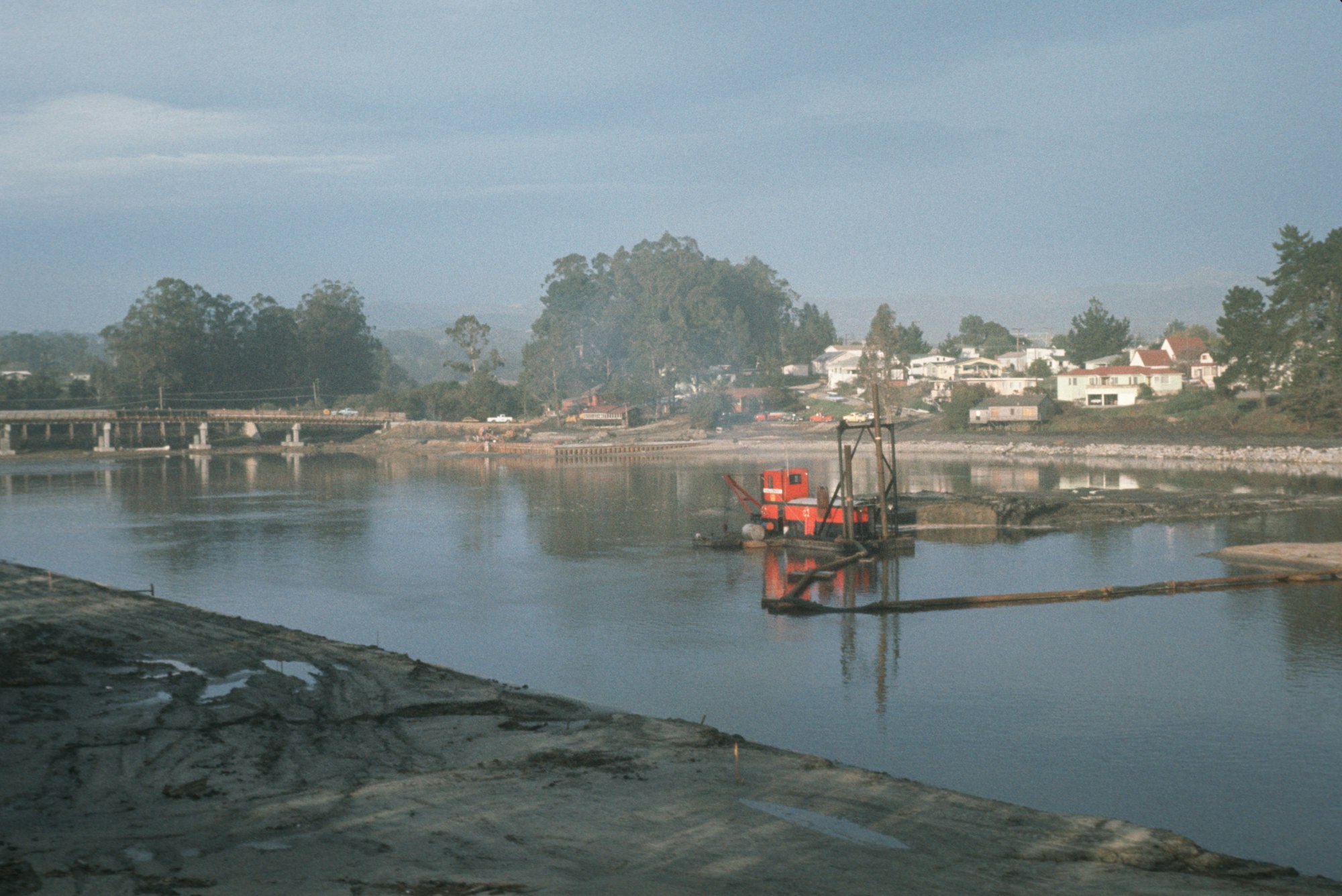 River dredging boat near a bridge and houses with trees in the background.