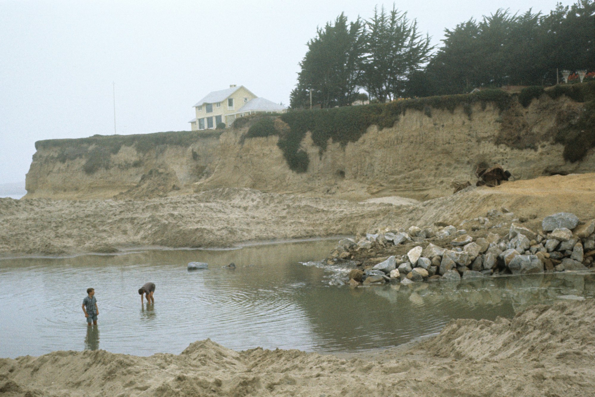 A coastal erosion scene with two people by a water pool, a damaged cliff, and a house on the edge.
