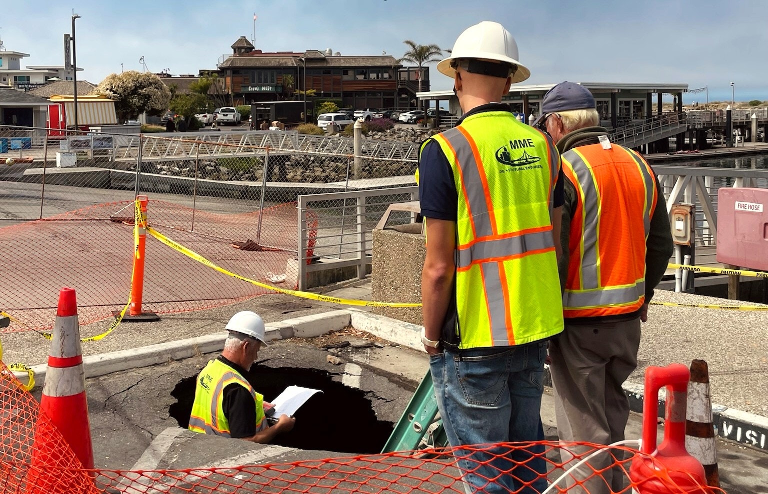 Construction workers in hard hats and vests inspect a hole on a street near waterfront, surrounded by safety cones and tape.