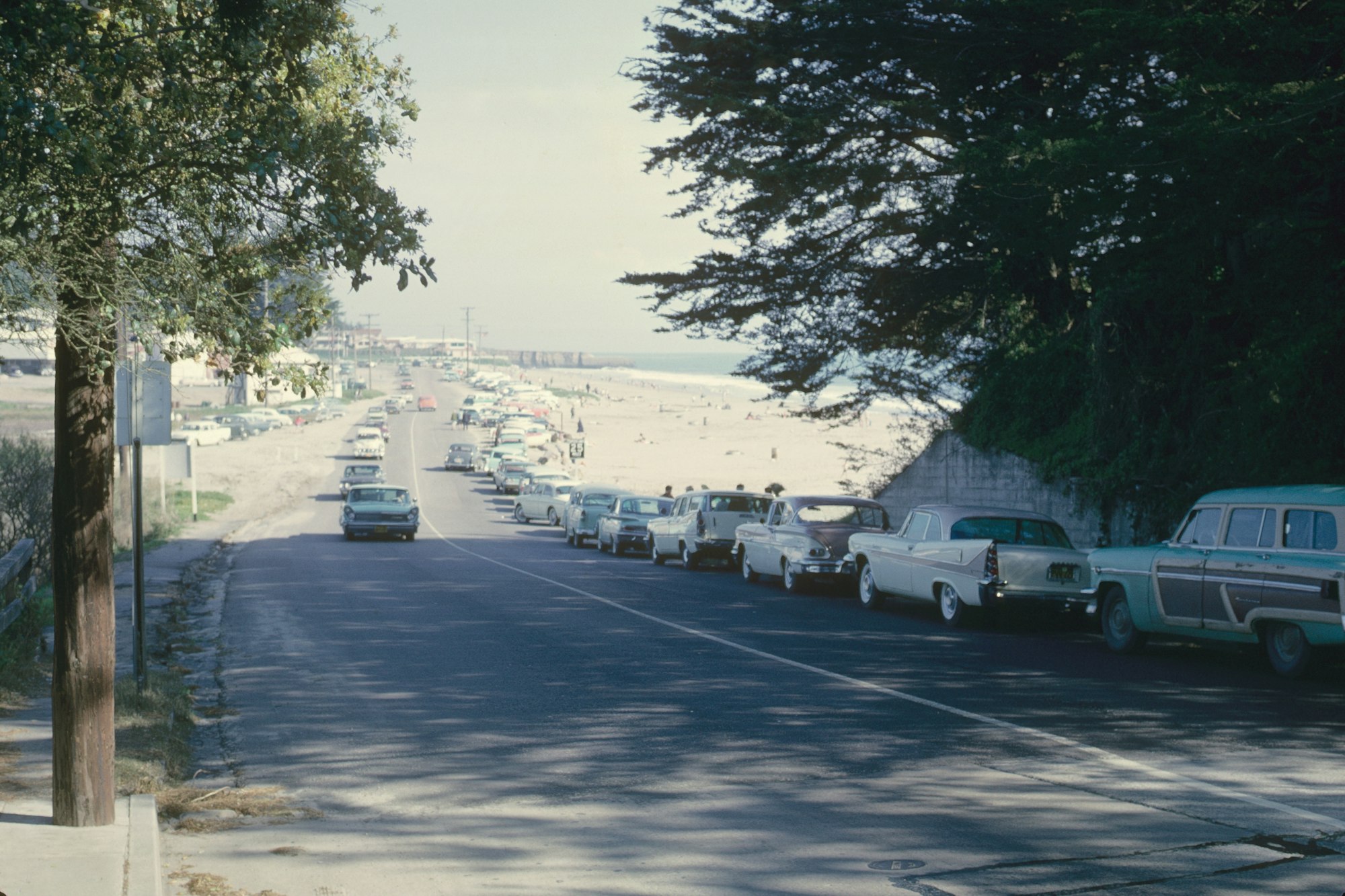 A vintage street view with classic cars parked along a beachside road, under a clear sky.