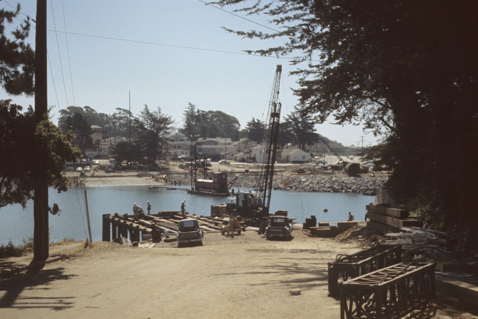 A waterfront construction site with a crane, boats, and vehicles, surrounded by trees and houses.