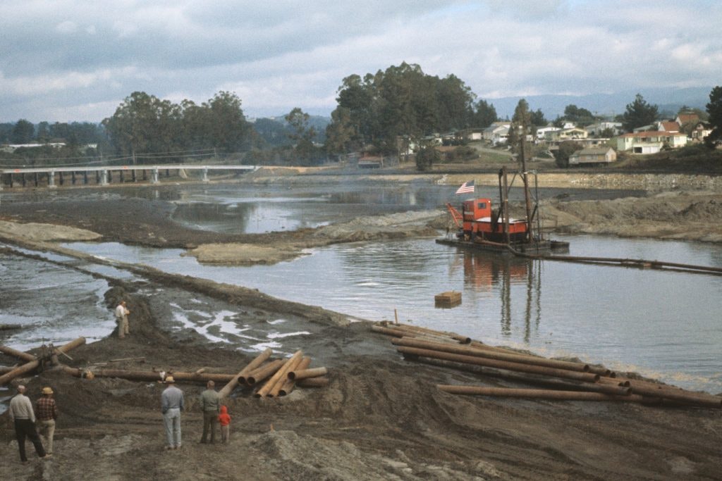 A dredging machine operates in a river, with people observing and wooden pipes nearby, amidst a scenic landscape of hills and homes.