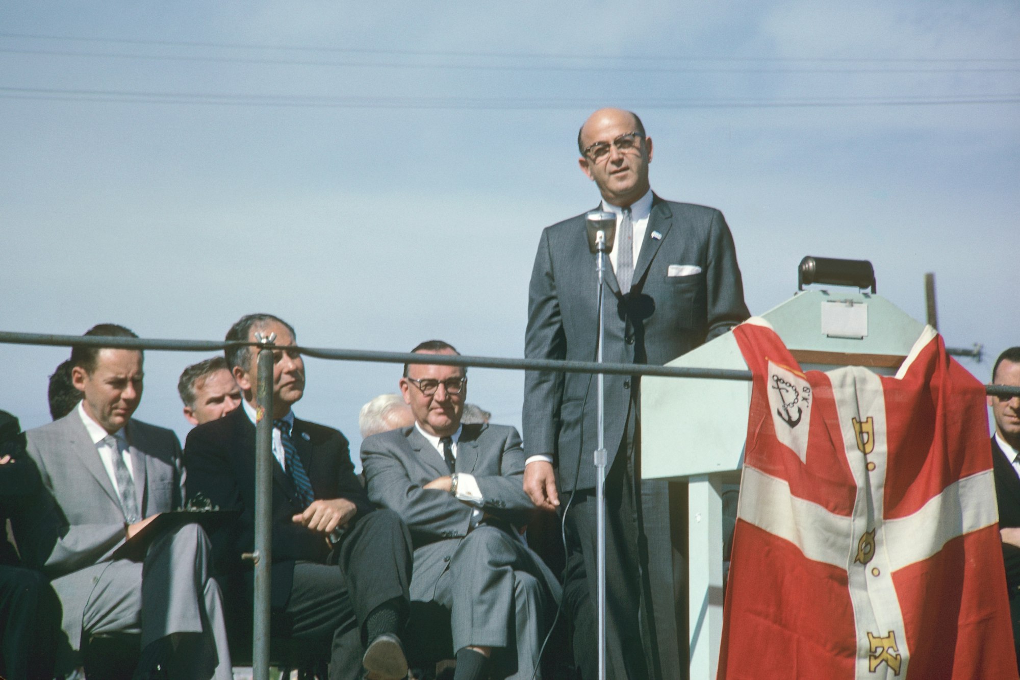 Men in suits seated at an event, one standing at a microphone, possibly giving a speech, with a ceremonial garment on display.