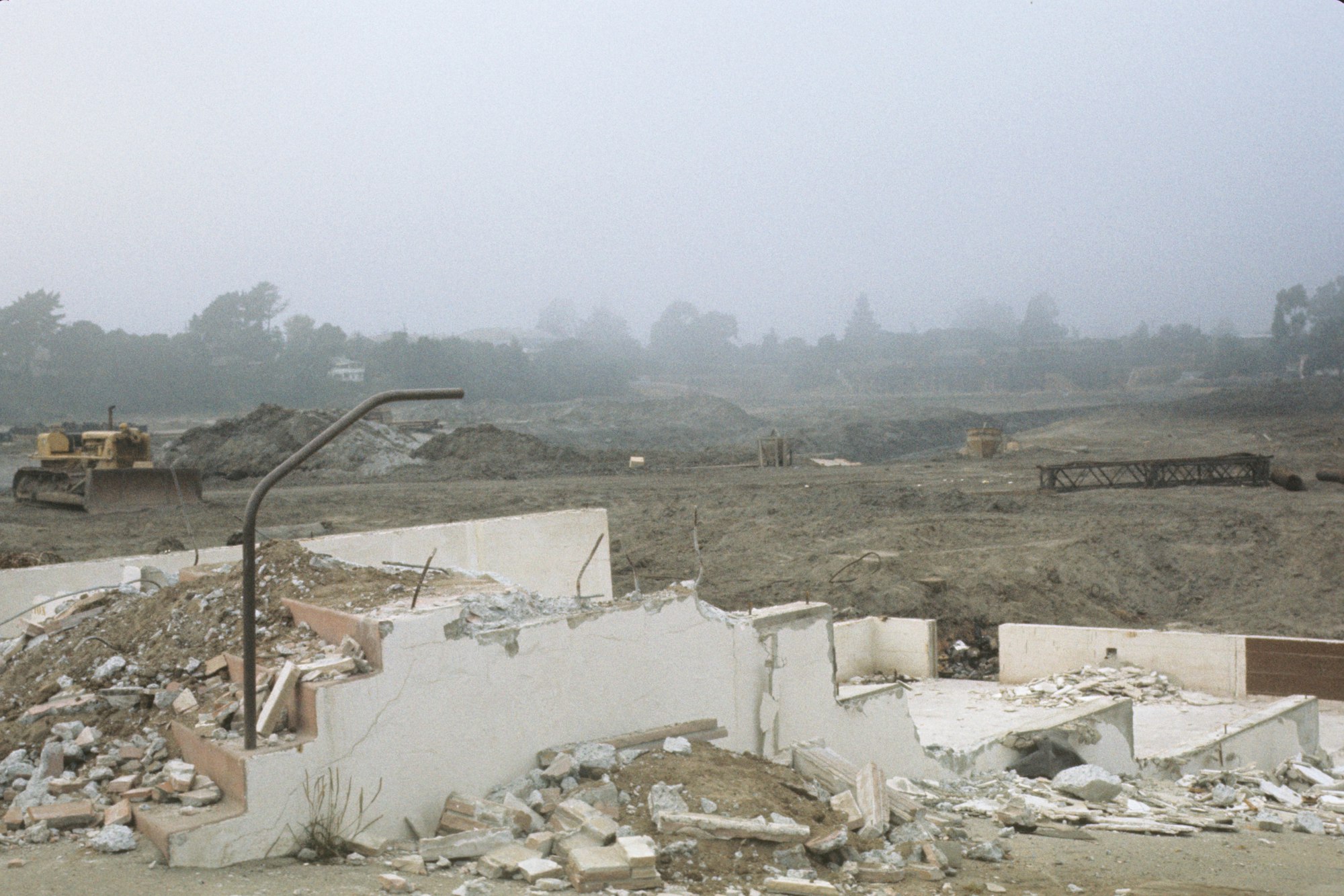 A demolished building with rubble, remnants of stairs, and construction vehicles in a foggy, desolate landscape.