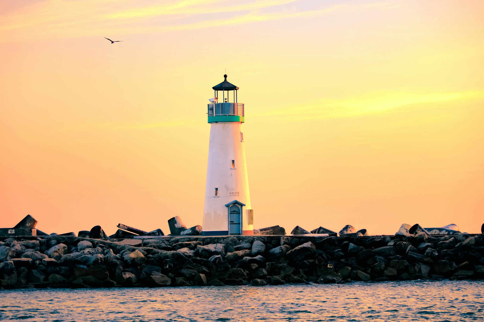 A lighthouse on a rocky shore with a bird flying above during a sunset.