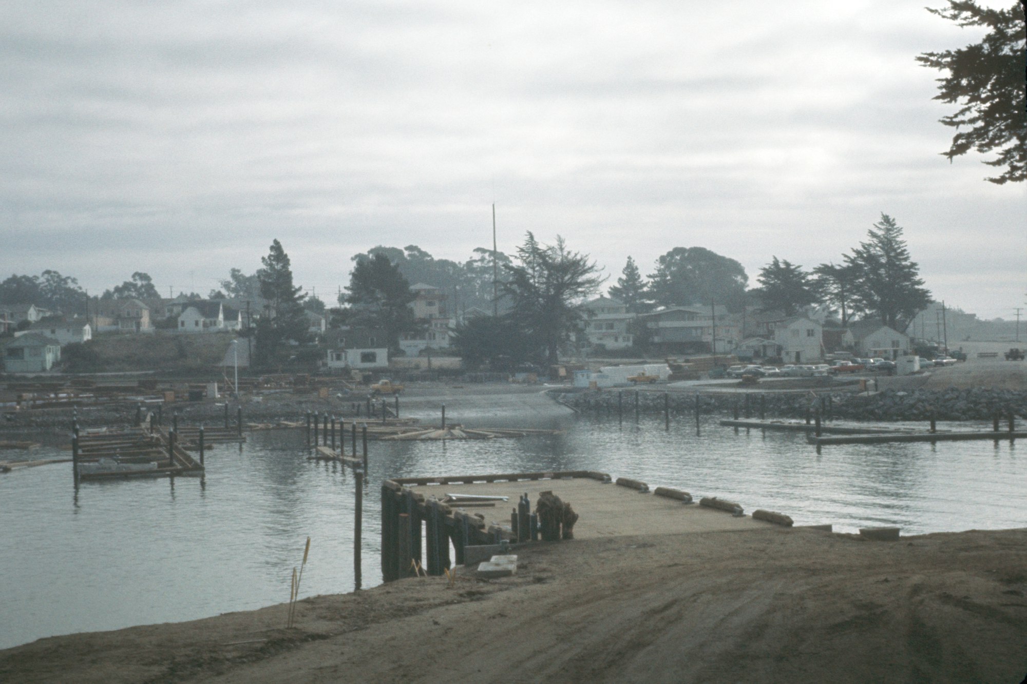 Waterfront construction area with pilings and equipment, houses in the background, overcast skies.