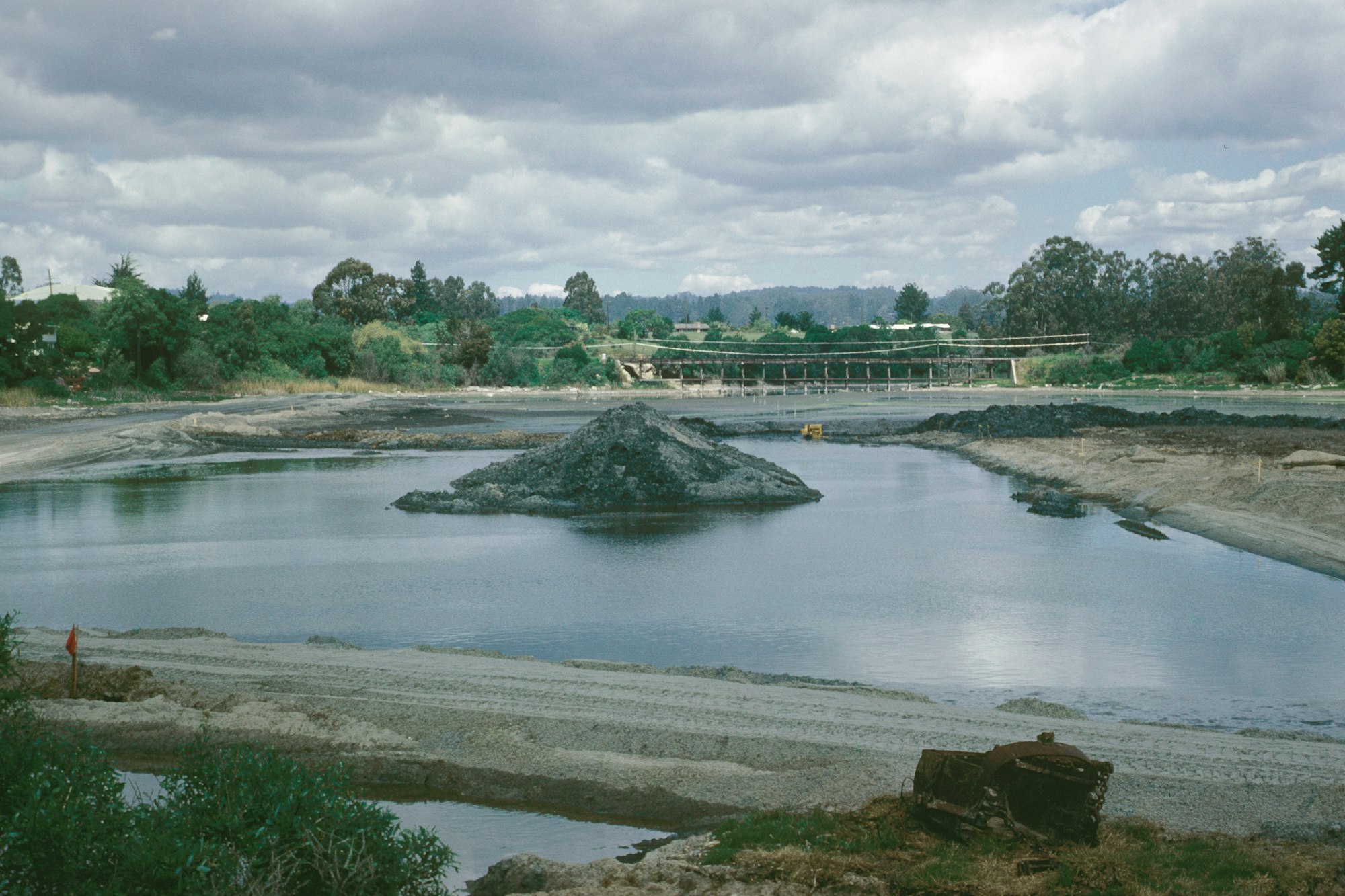 An excavation site with water, piles of earth, equipment, and a bridge in the background under a cloudy sky.
