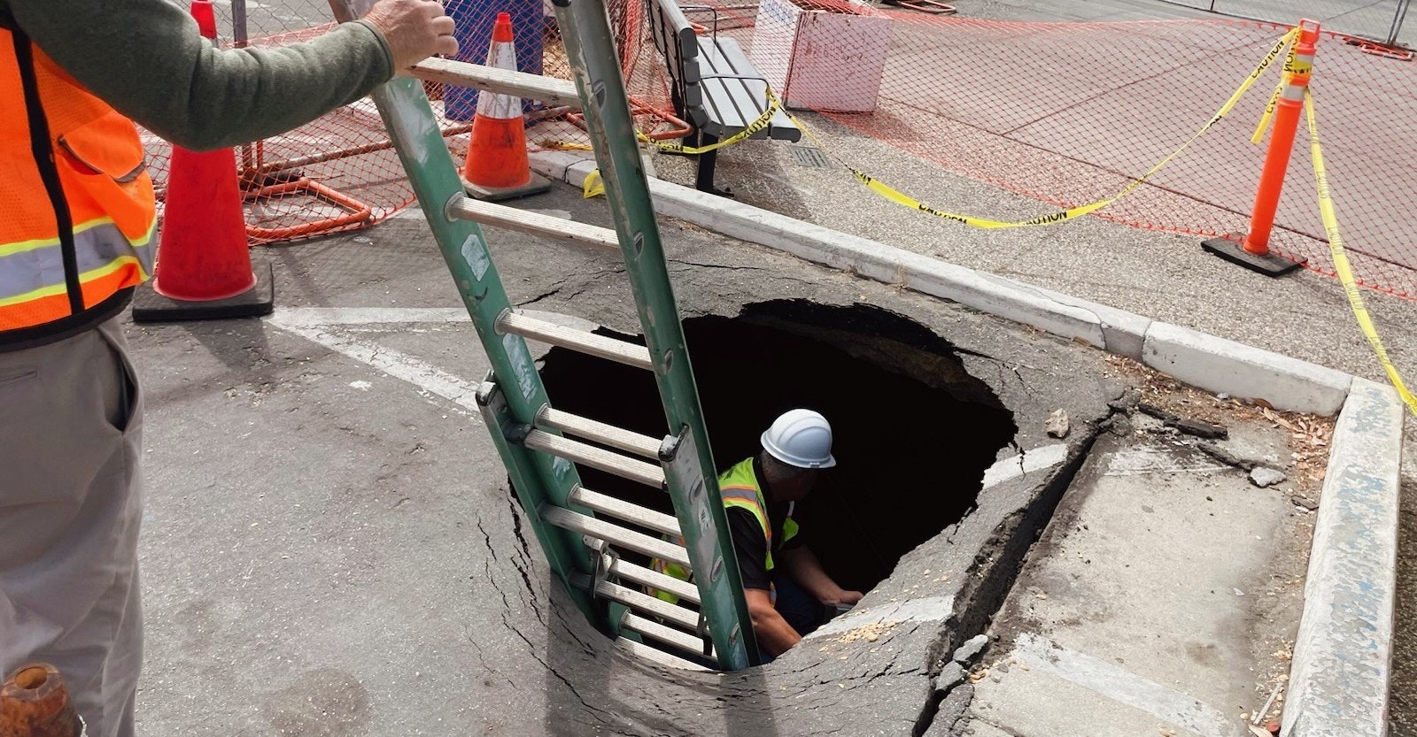 A worker in a hard hat descends a ladder into a large hole, surrounded by safety cones and caution tape.