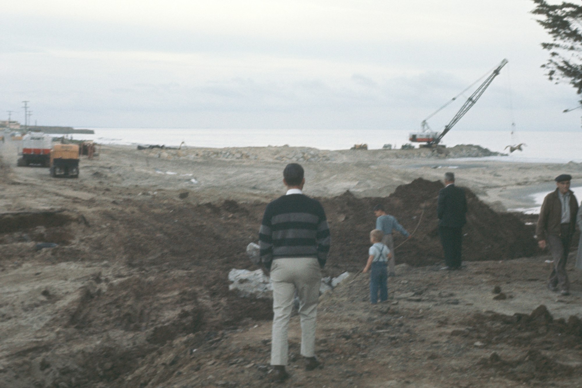 A construction site by the sea with a crane, vehicles, and several people including a child.