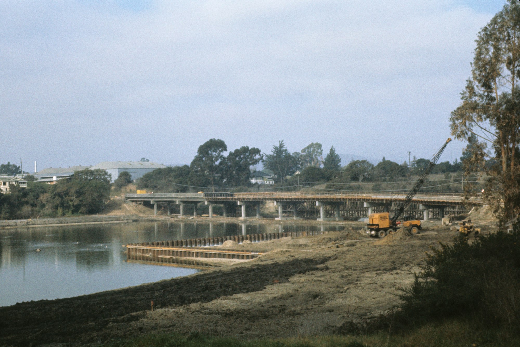 Bridge construction over water with crane and bulldozer, surrounded by greenery.