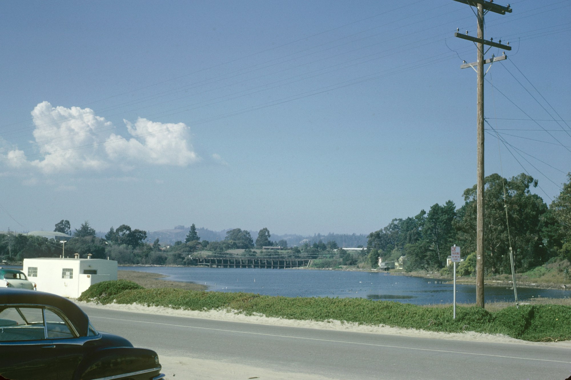 Scenic view of a river, bridge, trees, utility pole, and vintage car by the roadside under a blue sky with clouds.