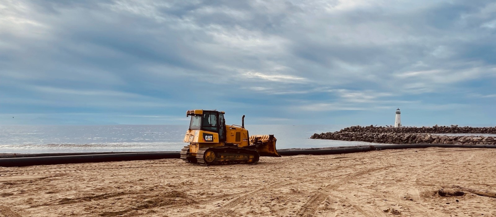 A bulldozer on a sandy beach, large pipe, rocky jetty, and distant lighthouse under a cloudy sky.