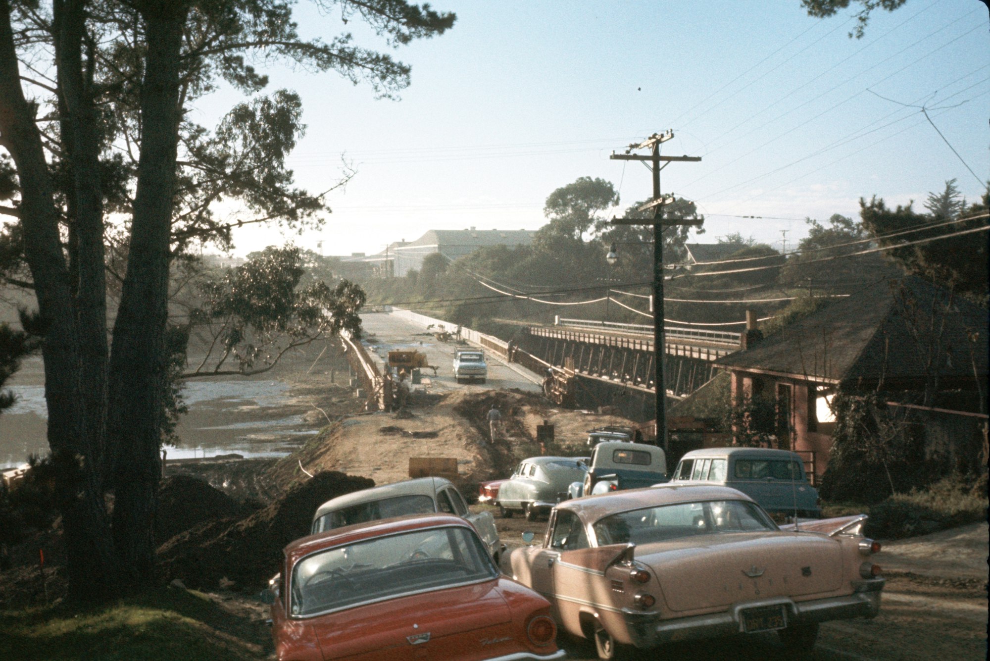 Vintage cars on a road near construction or road repair with trees and a bridge in the background.