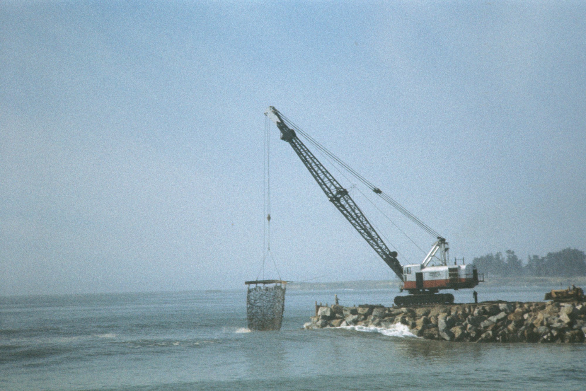 A crane lifting a large basket over a rocky coastline with the ocean in the background.