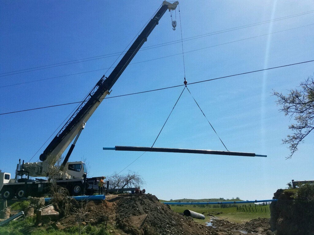 A crane is lifting a long object, possibly a pipe, in a construction area under clear blue skies.