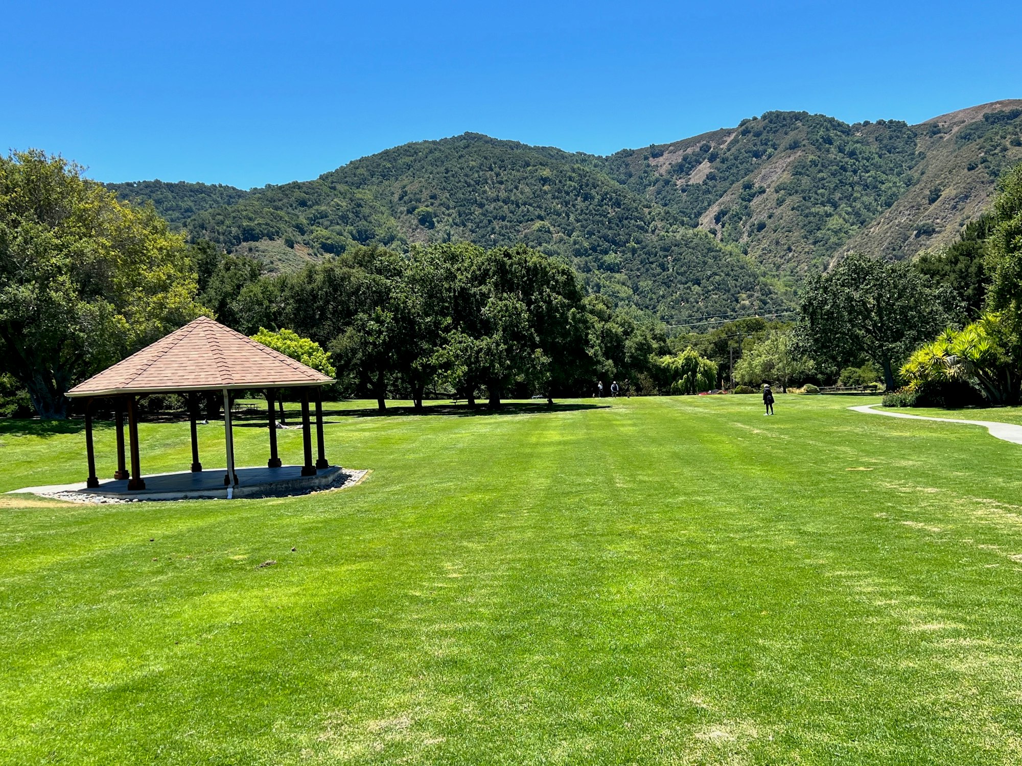 A gazebo on a large green field, with trees and hills under a clear blue sky.