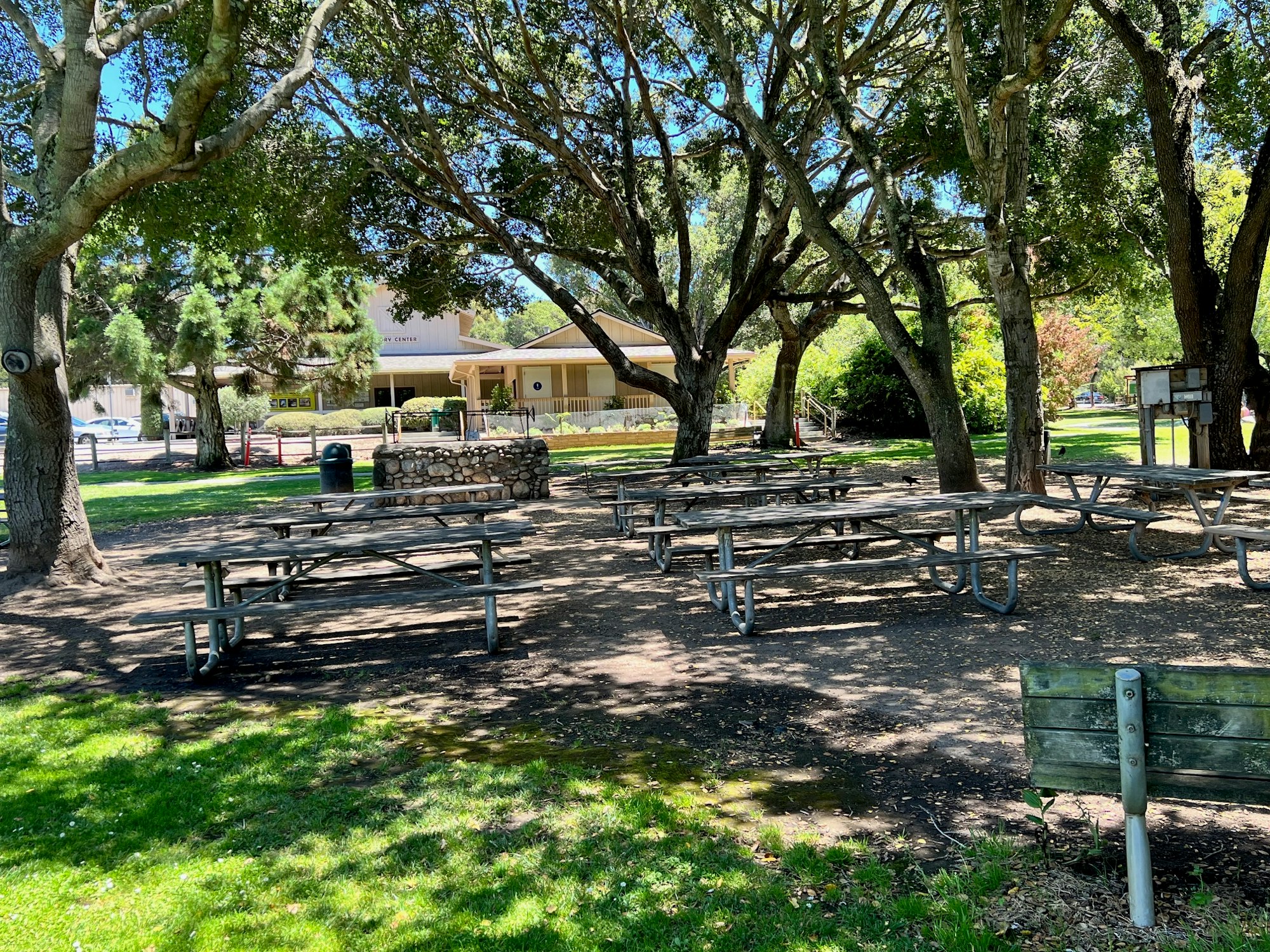 A park with picnic tables, trees, a stone structure, and a building in the background under a clear blue sky.