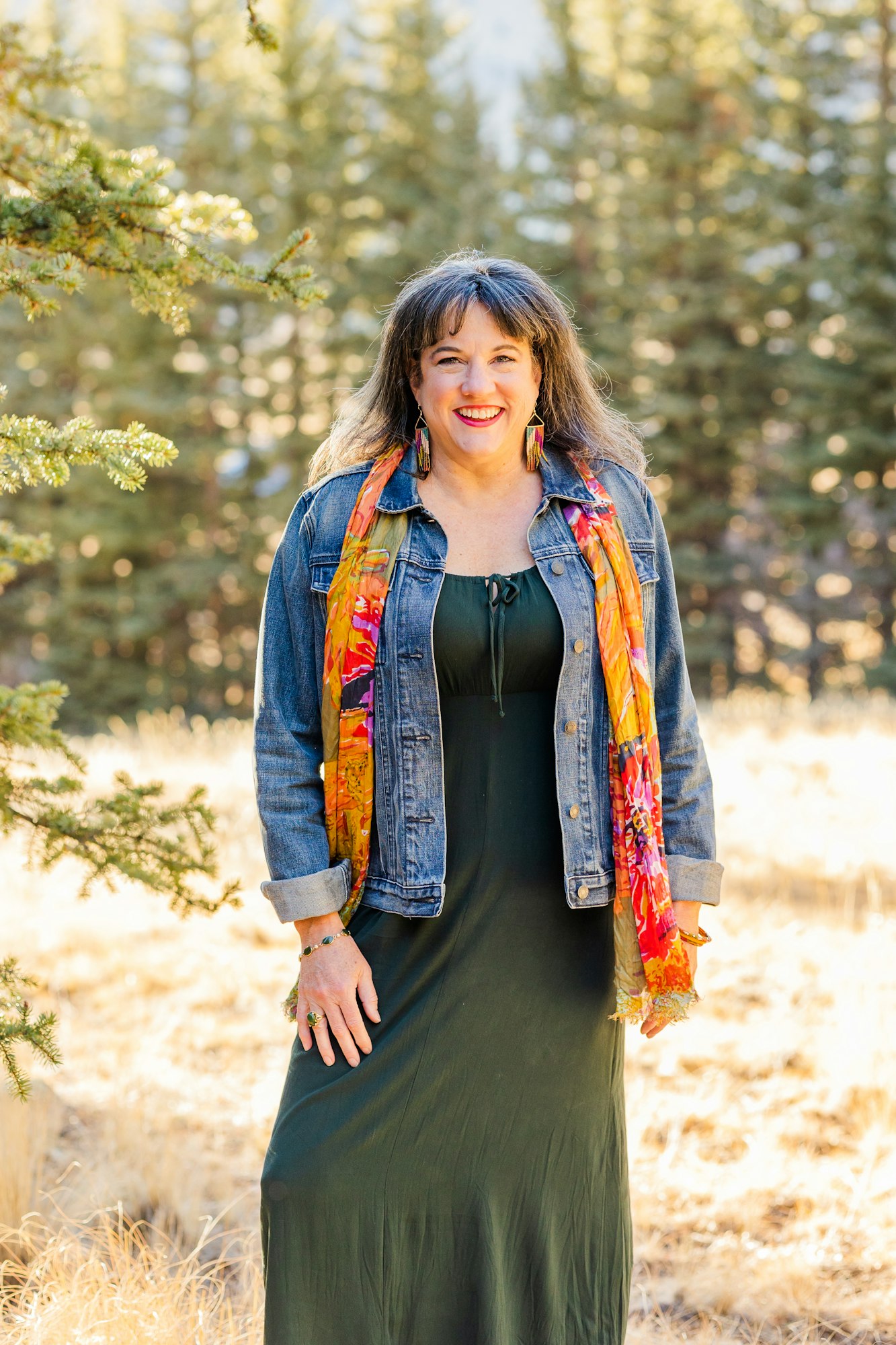 A smiling person outdoors wearing a denim jacket, green dress, and colorful scarf with trees in the background.